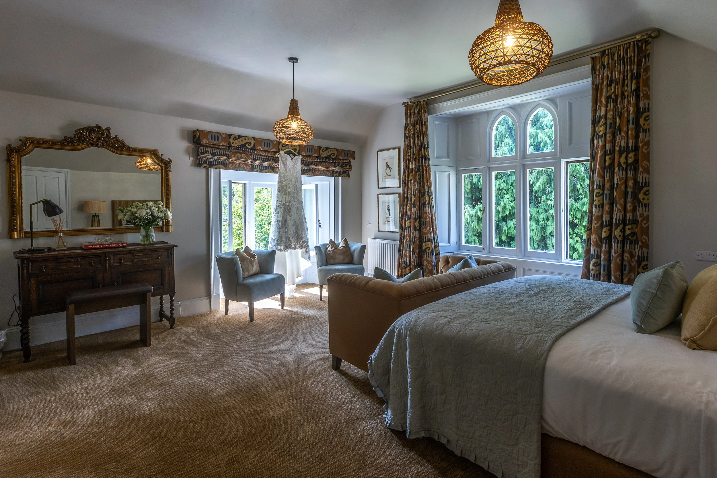 Spacious bedroom with a large bed, sitting area with two chairs, a wooden dresser with a large mirror, and big windows with patterned curtains overlooking greenery.
