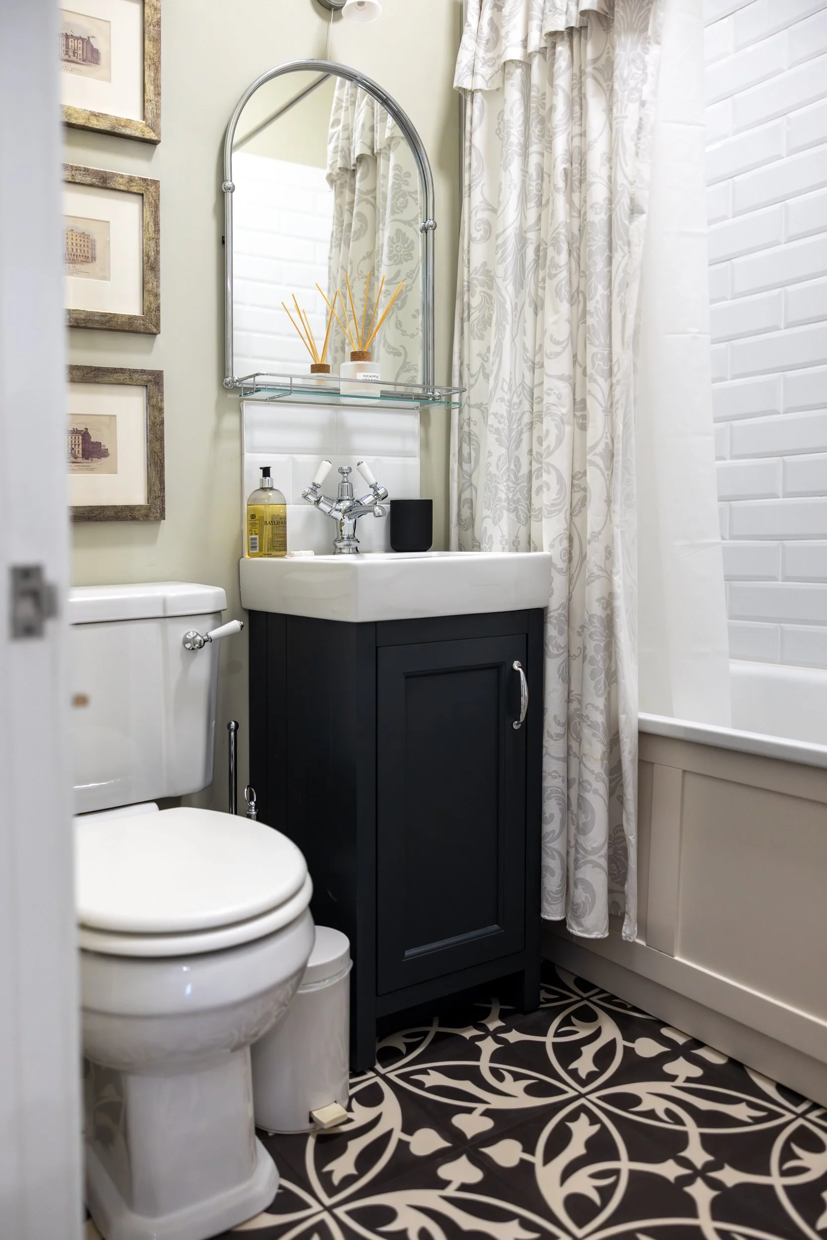 Small bathroom with a white toilet, dark vanity with a white sink, mirror, black soap dispenser, patterned black and white floor, white bathtub, and cream-colored floral curtains.