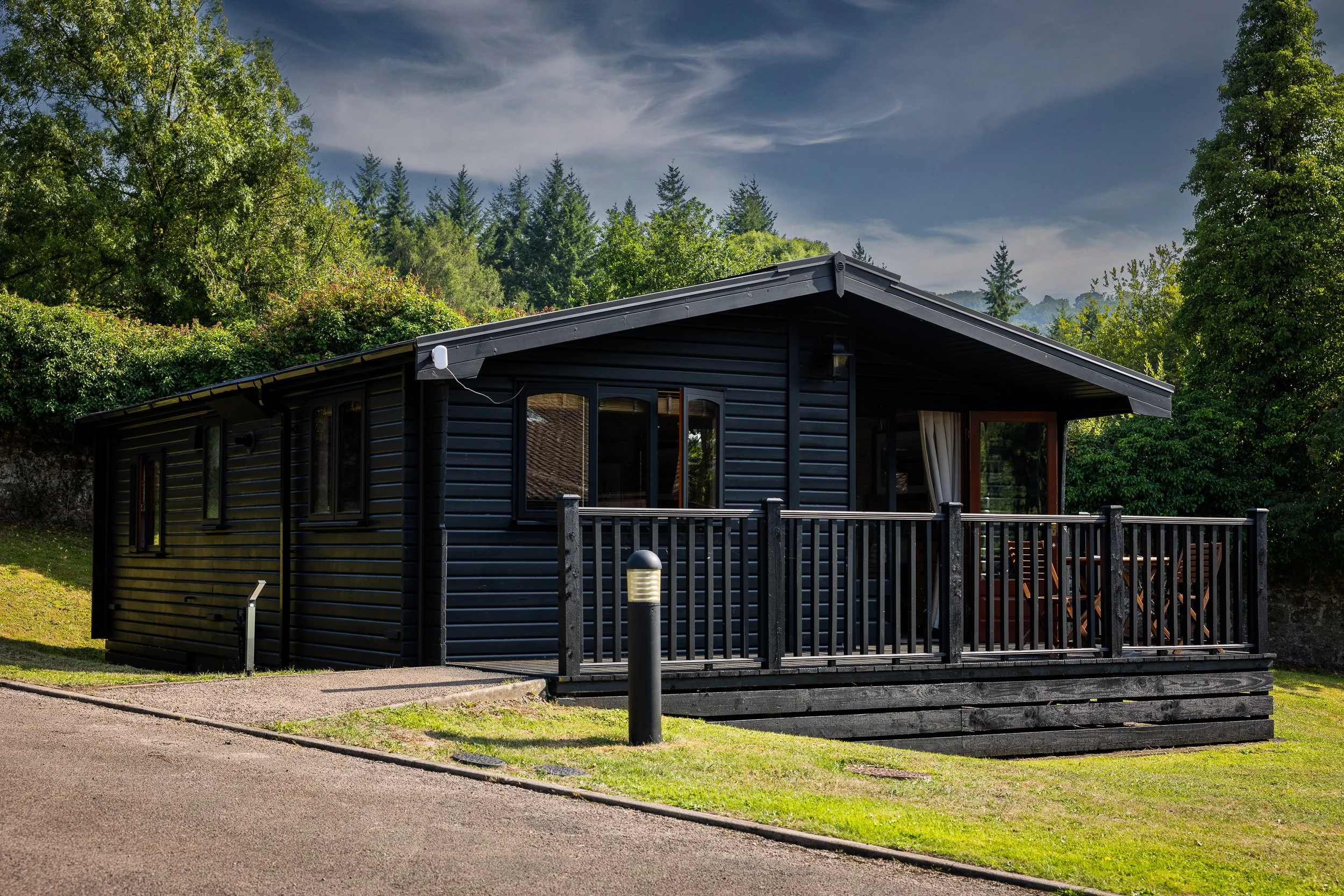 A black wooden cabin with a deck, surrounded by greenery and trees, under a partly cloudy sky.
