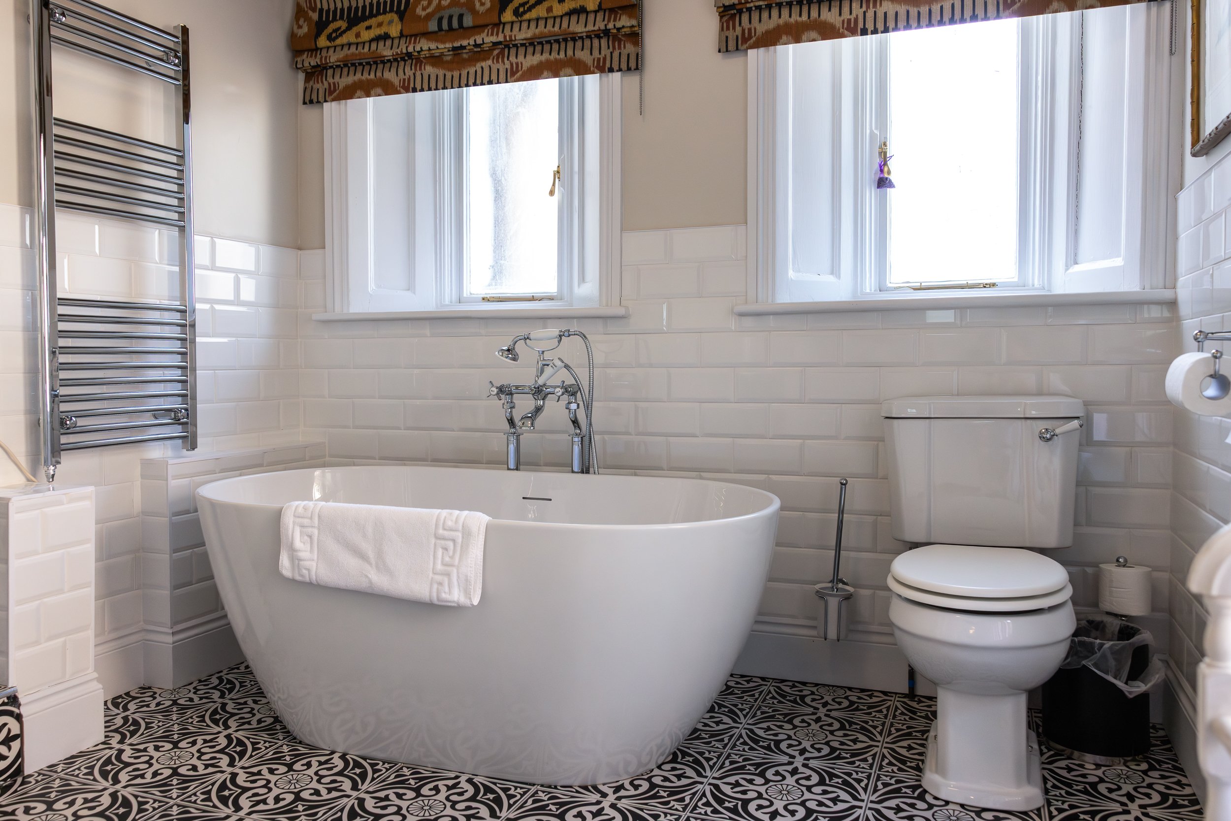 A bathroom with white brick tiles, a large white bathtub with a white towel hanging over the side, a toilet, two windows with white frames, patterned curtains, and black and white patterned floor tiles.