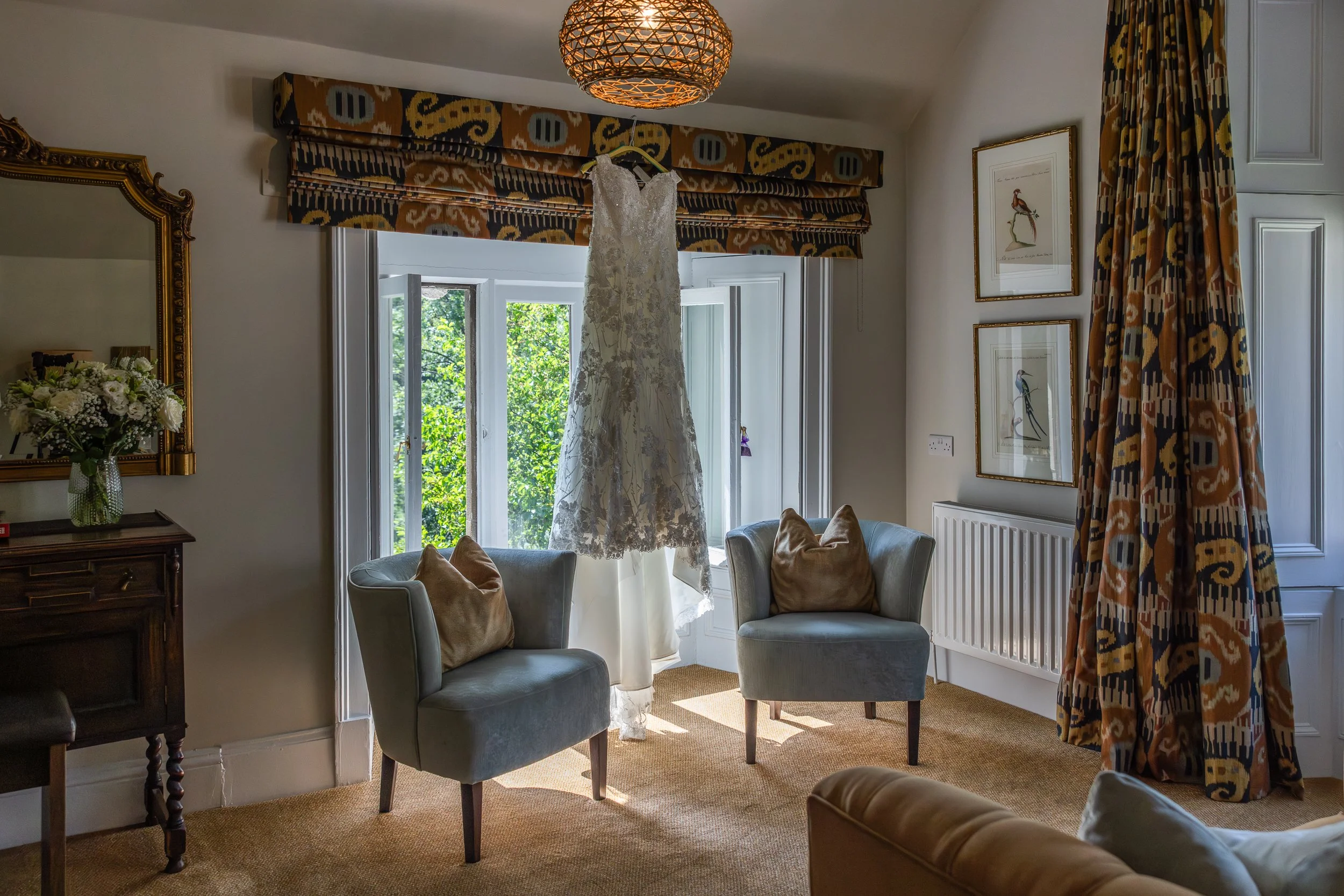 A wedding dress hanging in front of an open window in a living room with carpets, chairs, curtain, framed bird artwork, and a vase of flowers.