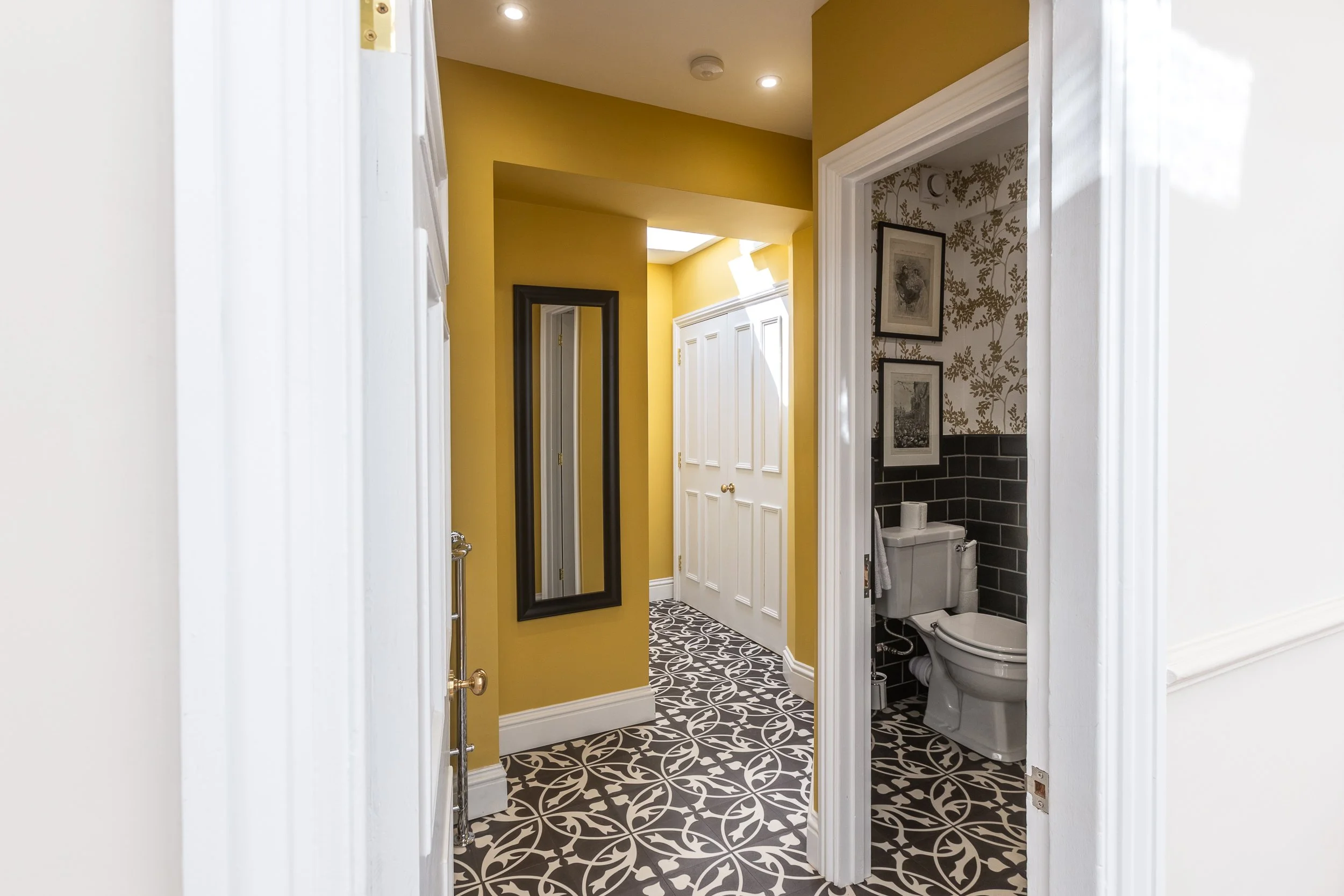 View of a hallway with a bathroom to the right, featuring yellow walls, a black-framed mirror, and black and white patterned flooring.