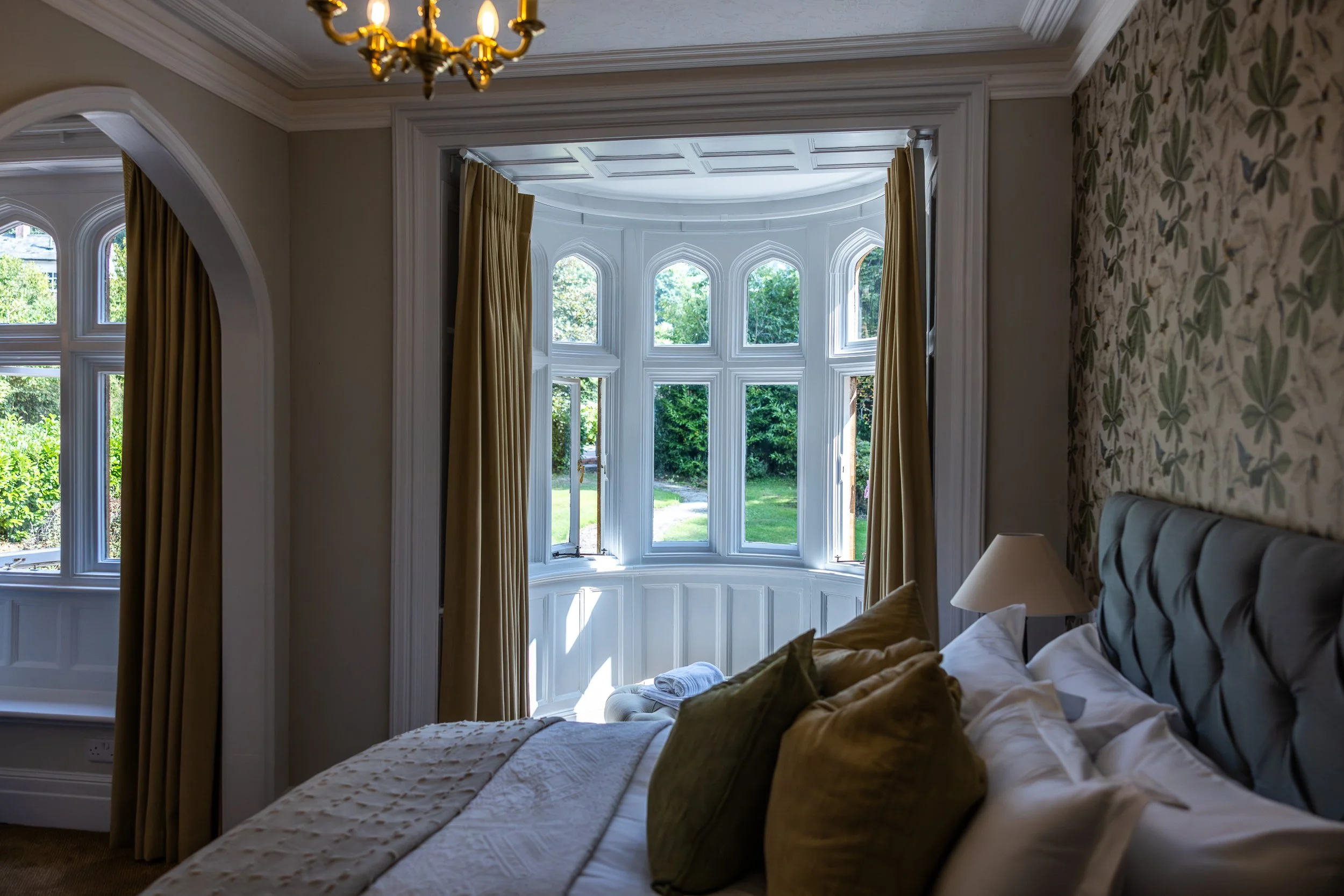 A bedroom with a large bay window, beige curtains, a tufted headboard, a white bedspread, and a bedside lamp, with sunlight coming through the windows showing a green yard outside.