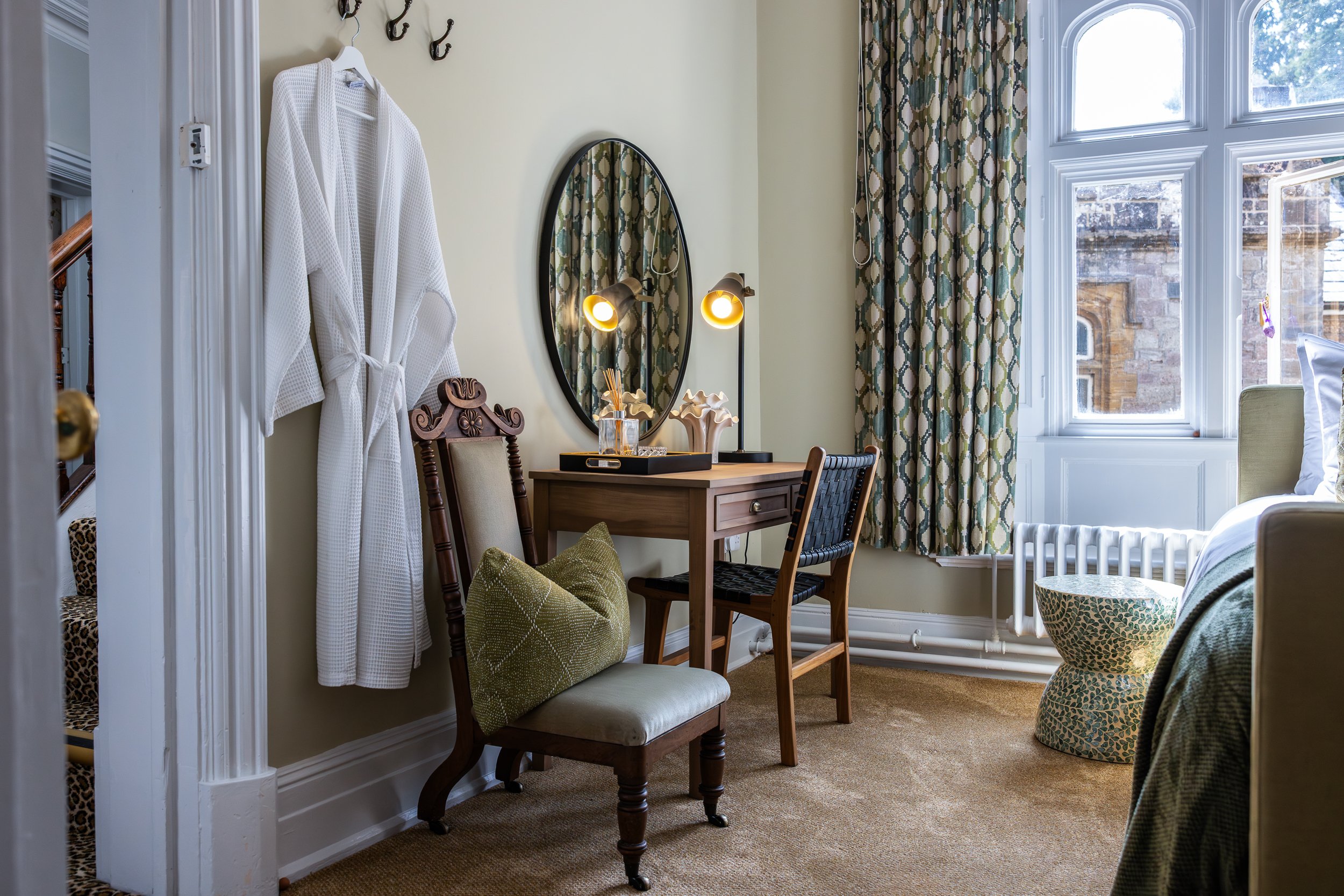 A cozy bedroom corner with a wooden desk, two chairs, a mirror, and a window with patterned curtains. A white bathrobe hangs on the wall.