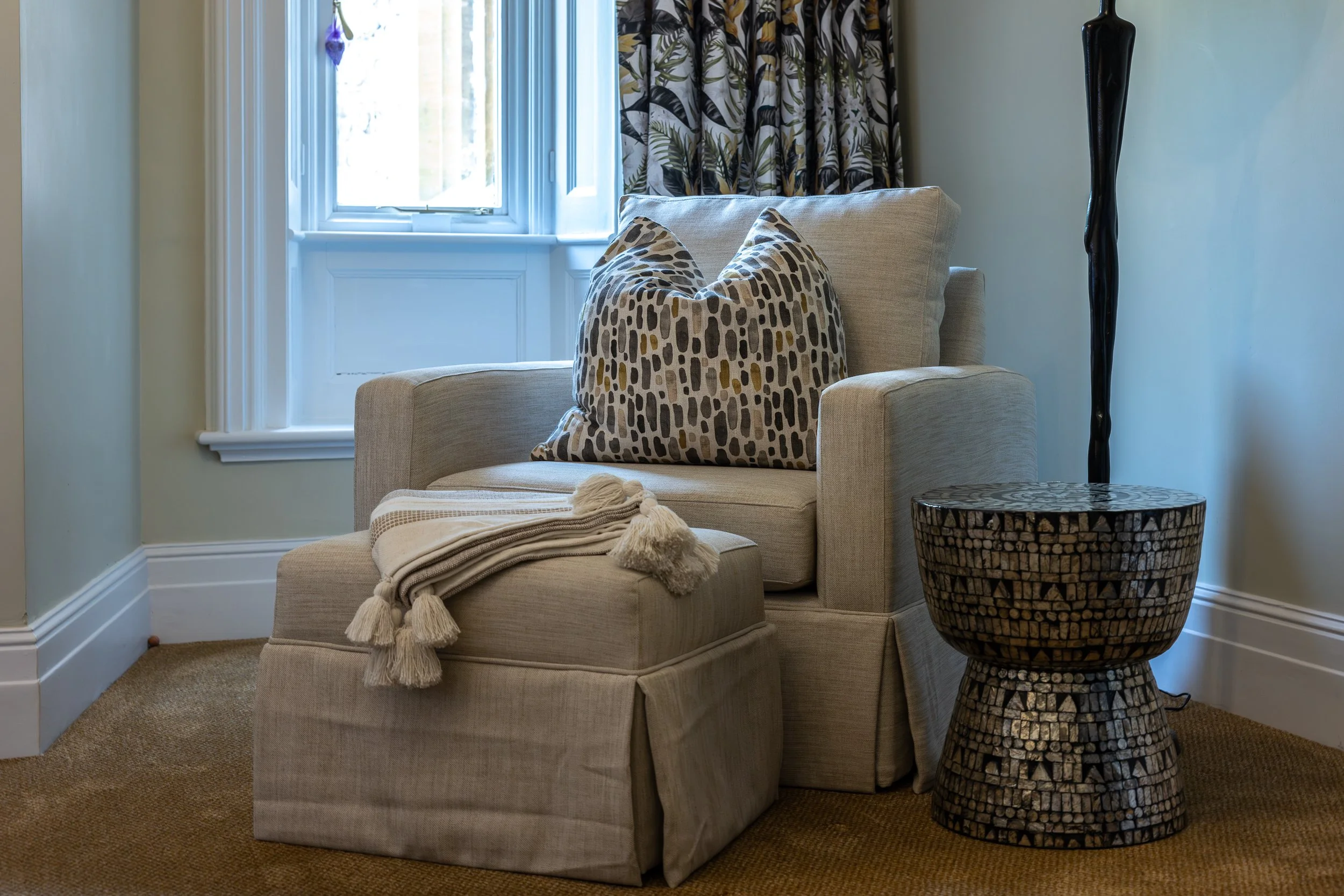 A beige armchair with a leopard print throw pillow, a throw blanket, and an ottoman, next to a patterned side table and a black floor lamp, near a window with patterned curtains.