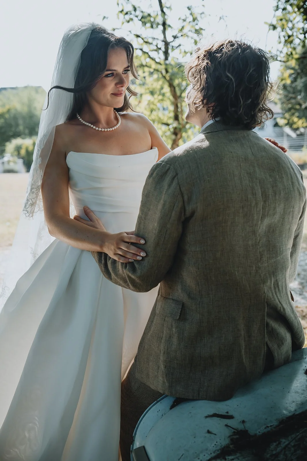 A bride with long dark hair, wearing a strapless white wedding dress, a pearl necklace, and a veil, looking at a man with curly hair wearing a brown suit, sitting on a car, outdoors with trees in the background.