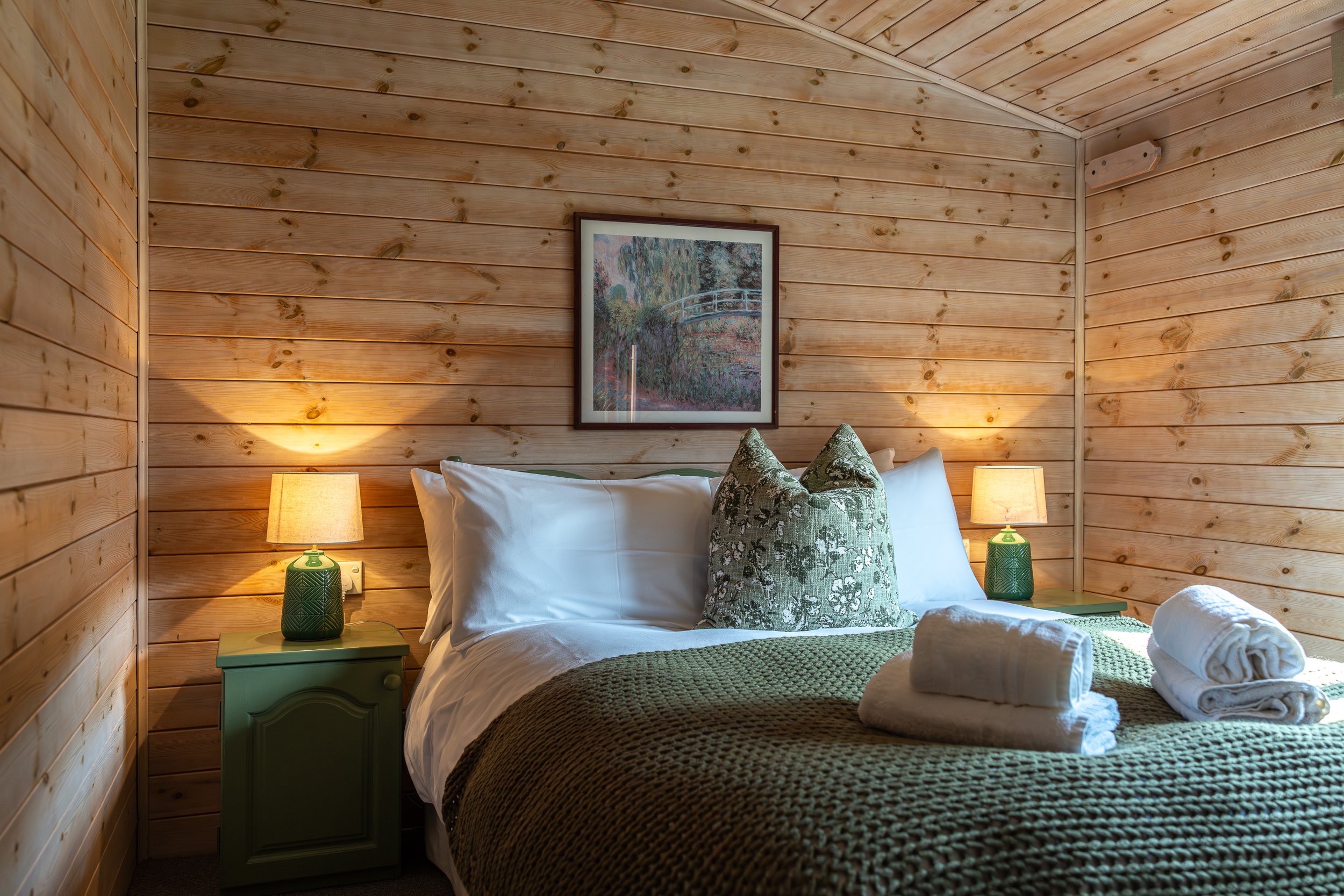 A cozy bedroom with wooden panel walls, a double bed with white linens, green knitted bedspread, decorative pillows, and two green nightstands with matching lamps, framed artwork hanging above the bed, and folded towels on the bed.