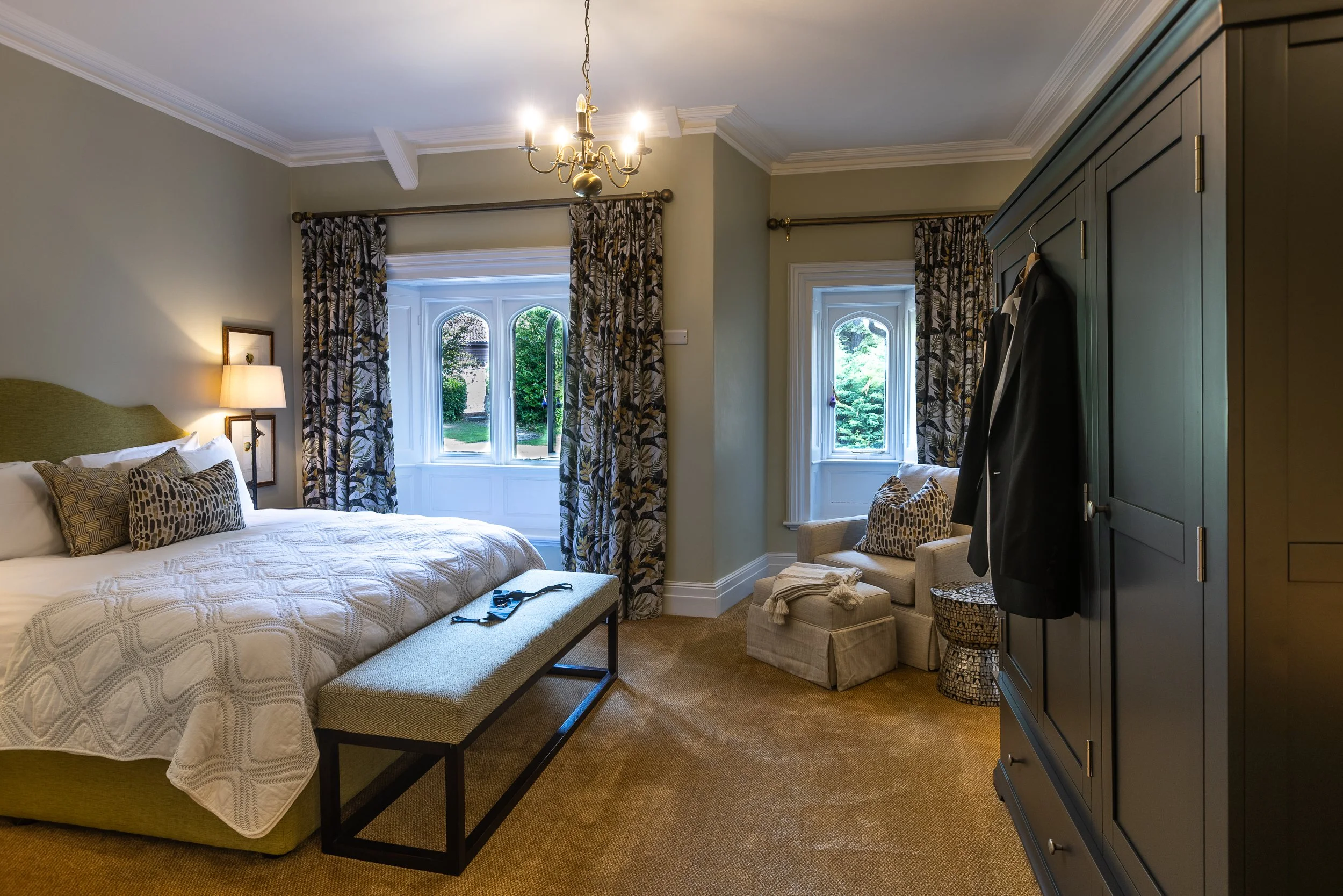 A bedroom with a large bed, green upholstered headboard, patterned pillows, a bench at the foot of the bed, and a side table with a lamp. There is a chair with a pillow, a wardrobe with hanging clothes, and windows with floral curtains overlooking a 