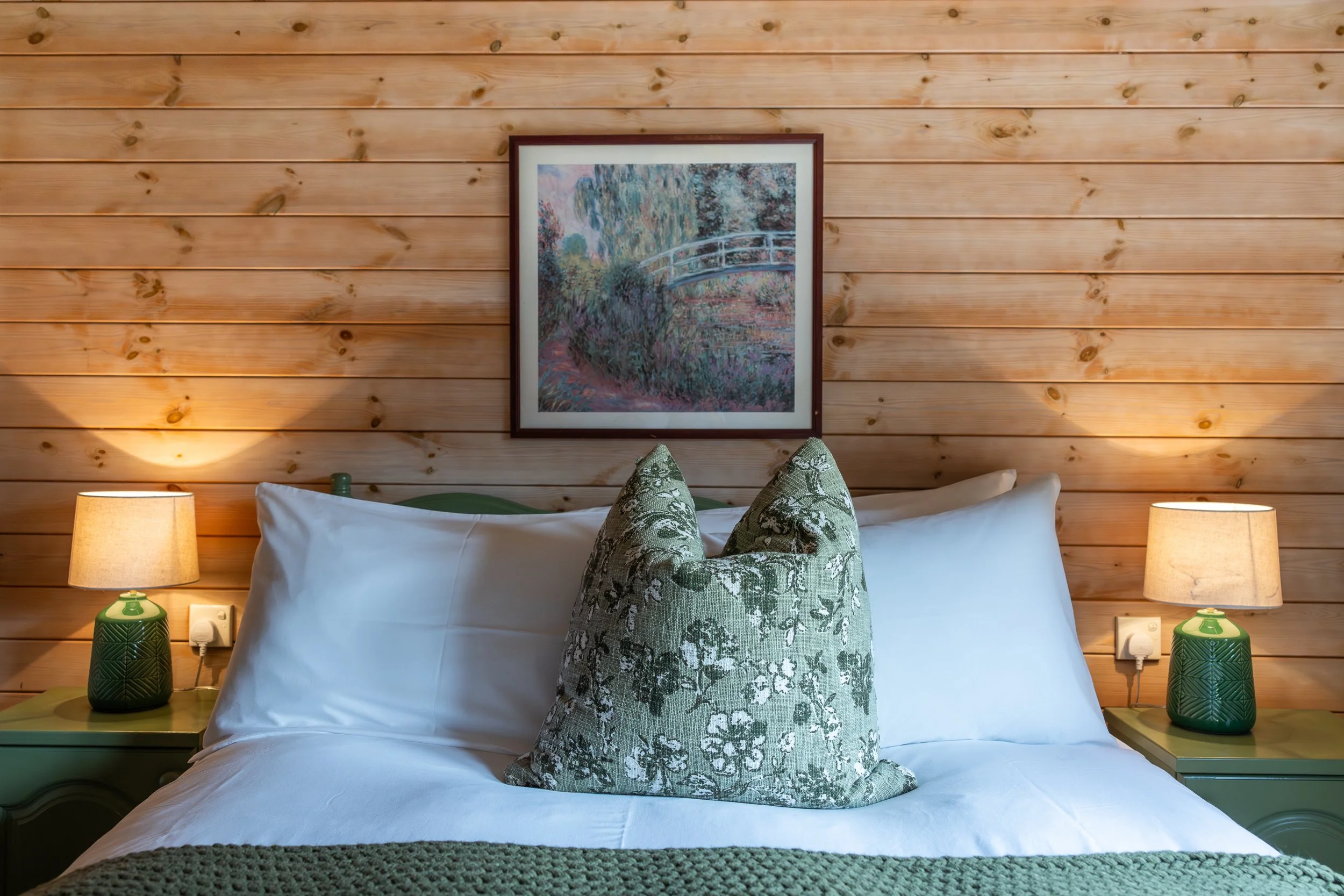 A cozy bedroom with a wooden wall, white bed with decorative green pillow, two matching green lamps, and a framed painting of a bridge and garden scene above the bed.