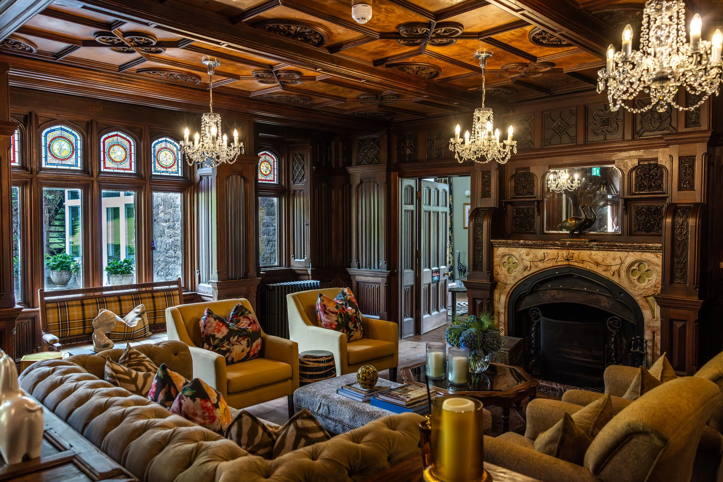 Living room with wood-paneled walls, stained glass windows, chandeliers, and vintage furniture, including tufted sofas, armchairs, and a marble fireplace with decorative elements and sculptures.