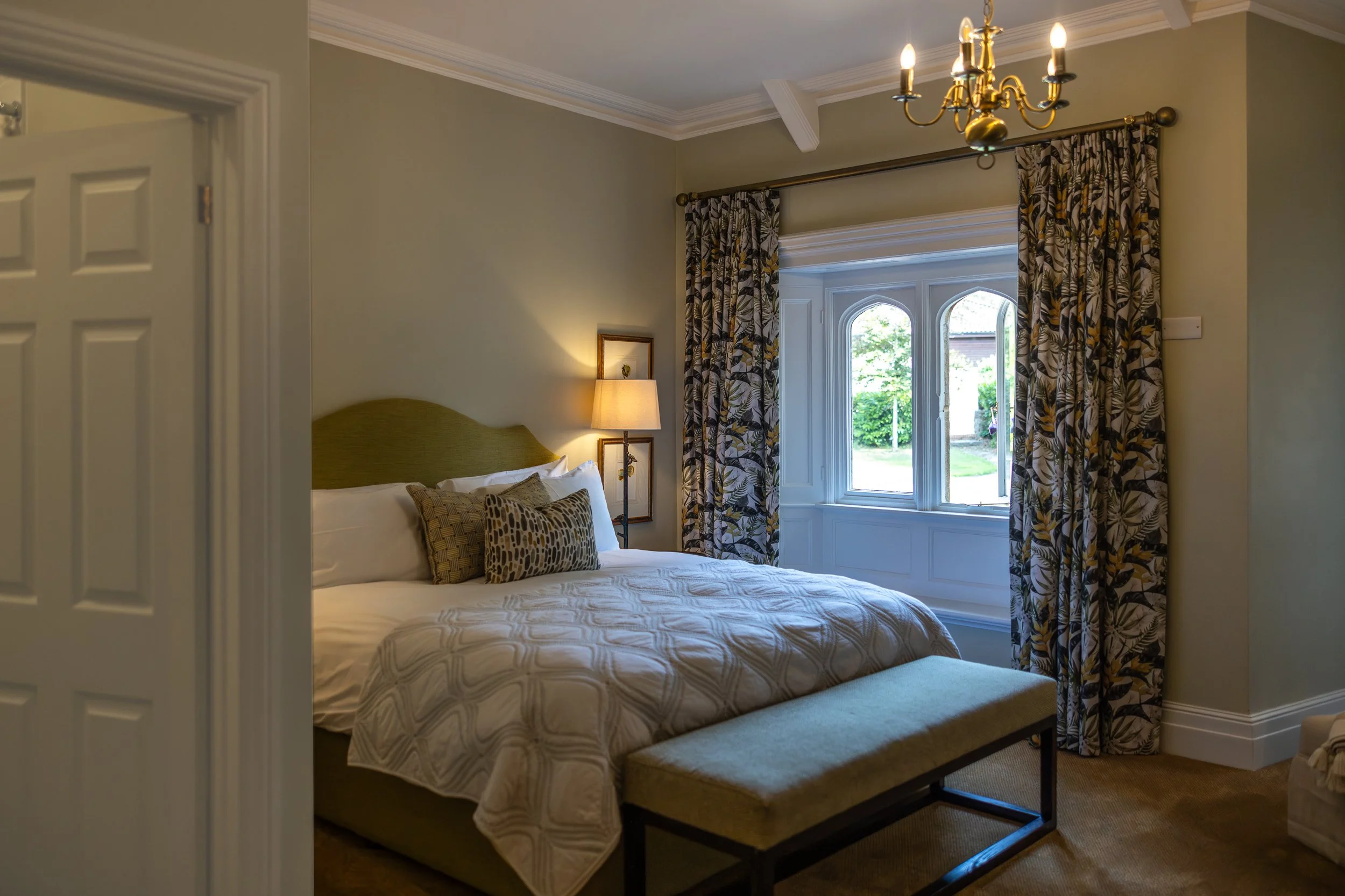 A cozy bedroom with a large bed, decorative pillows, a small bench at the foot, and a window with patterned curtains allowing sunlight in.