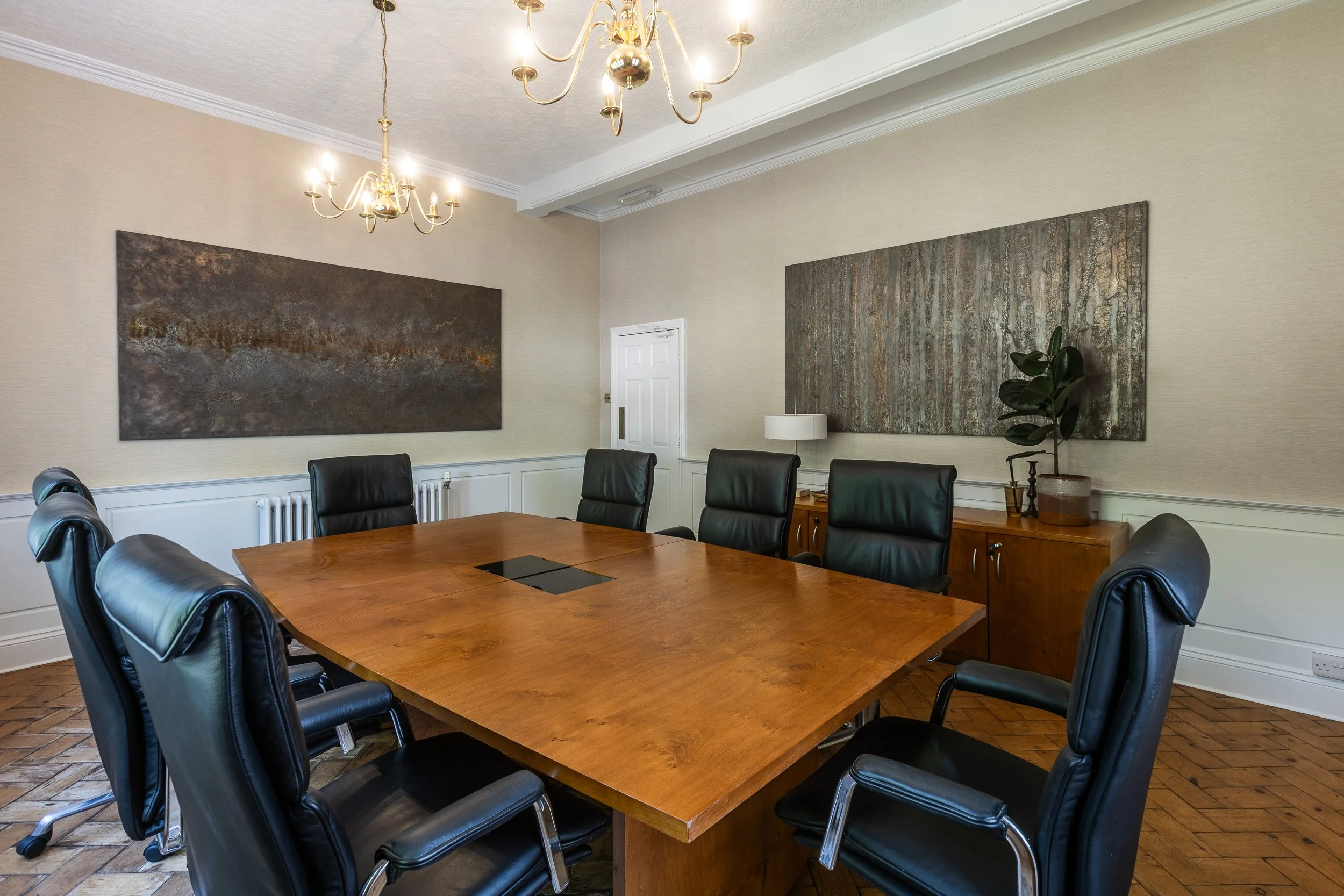 Empty conference room with a large wooden table surrounded by black leather chairs, hanging chandeliers, artwork on the beige walls, and a side cabinet with a potted plant.