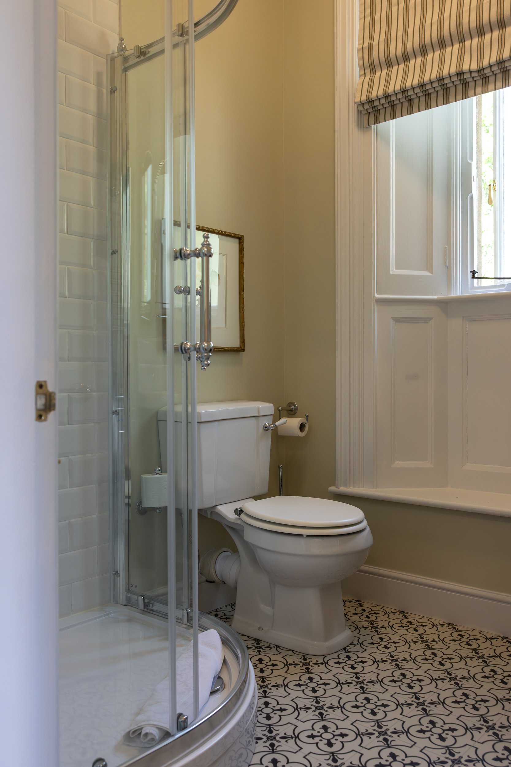 A small bathroom with a corner shower enclosure, a white toilet with a roll of toilet paper on the holder, and a window with a striped beige and white Roman shade. The walls are painted light beige, and the floor has black and white patterned tiles.