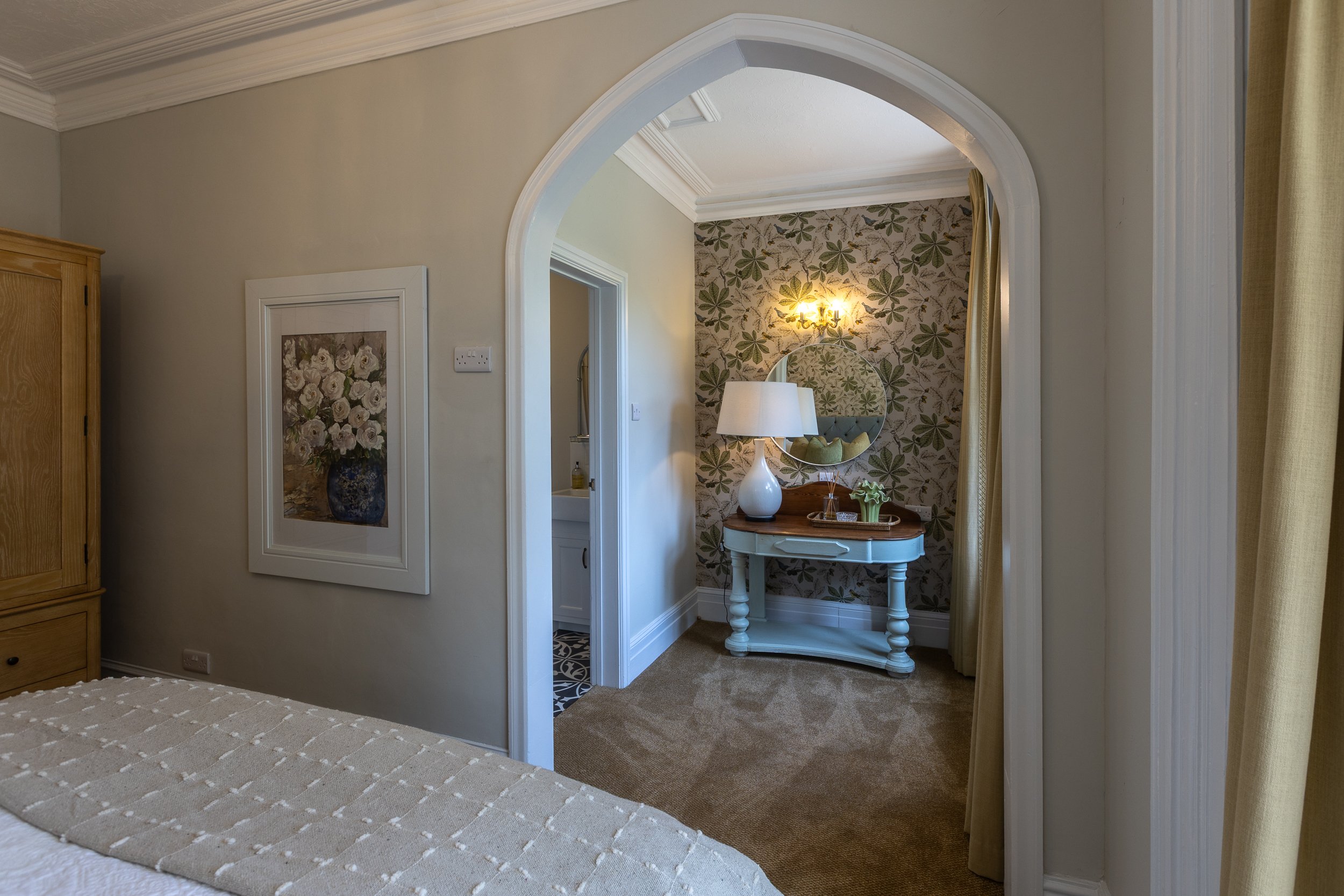 View of a bedroom with an arched doorway leading to a small alcove with a table, lamp, and mirror, and a bedroom wall with a flower-patterned wallpaper.