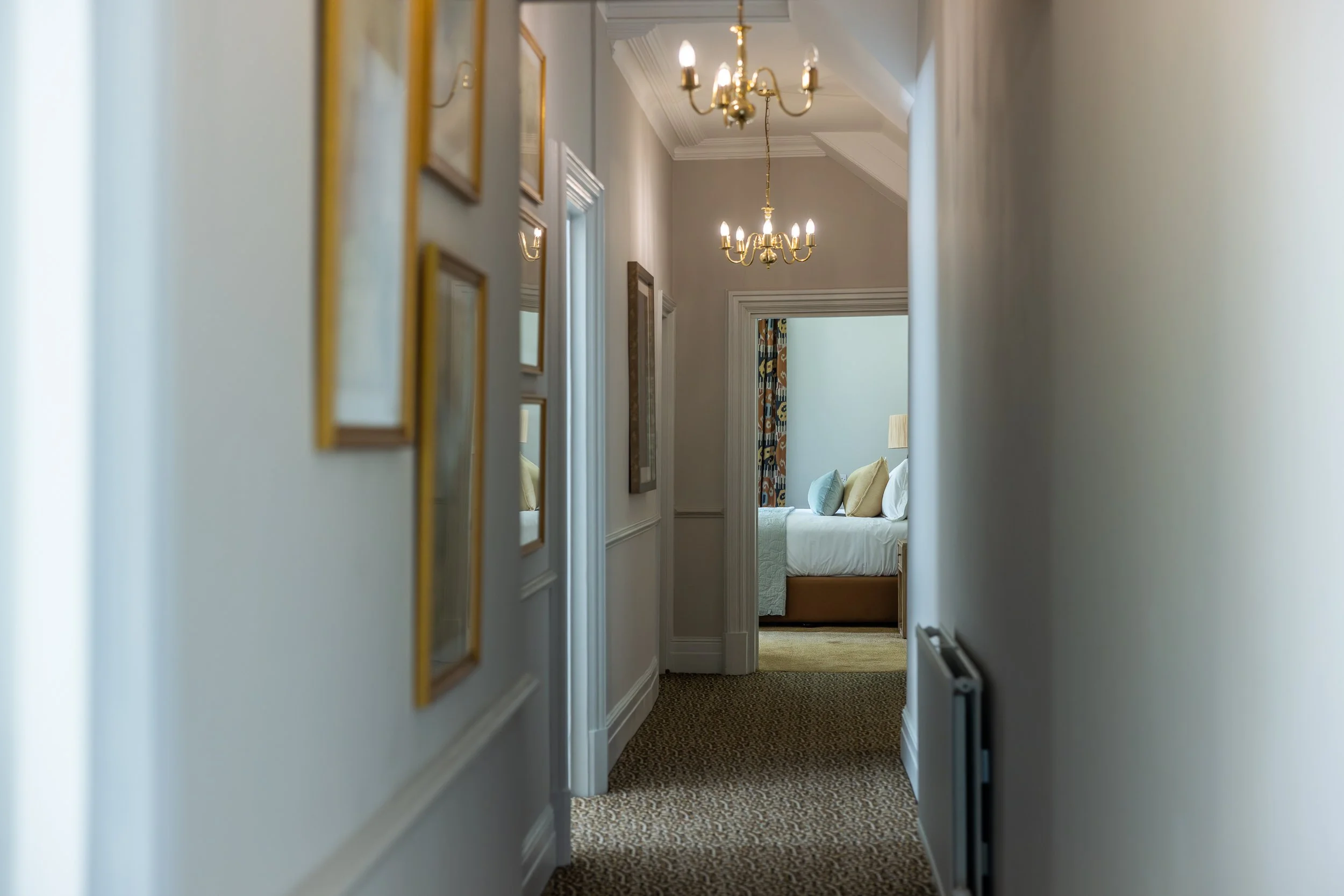 A narrow hallway in a hotel or home with framed pictures on the left wall, leading to a bedroom with a bed, pillows, and colorful curtains.