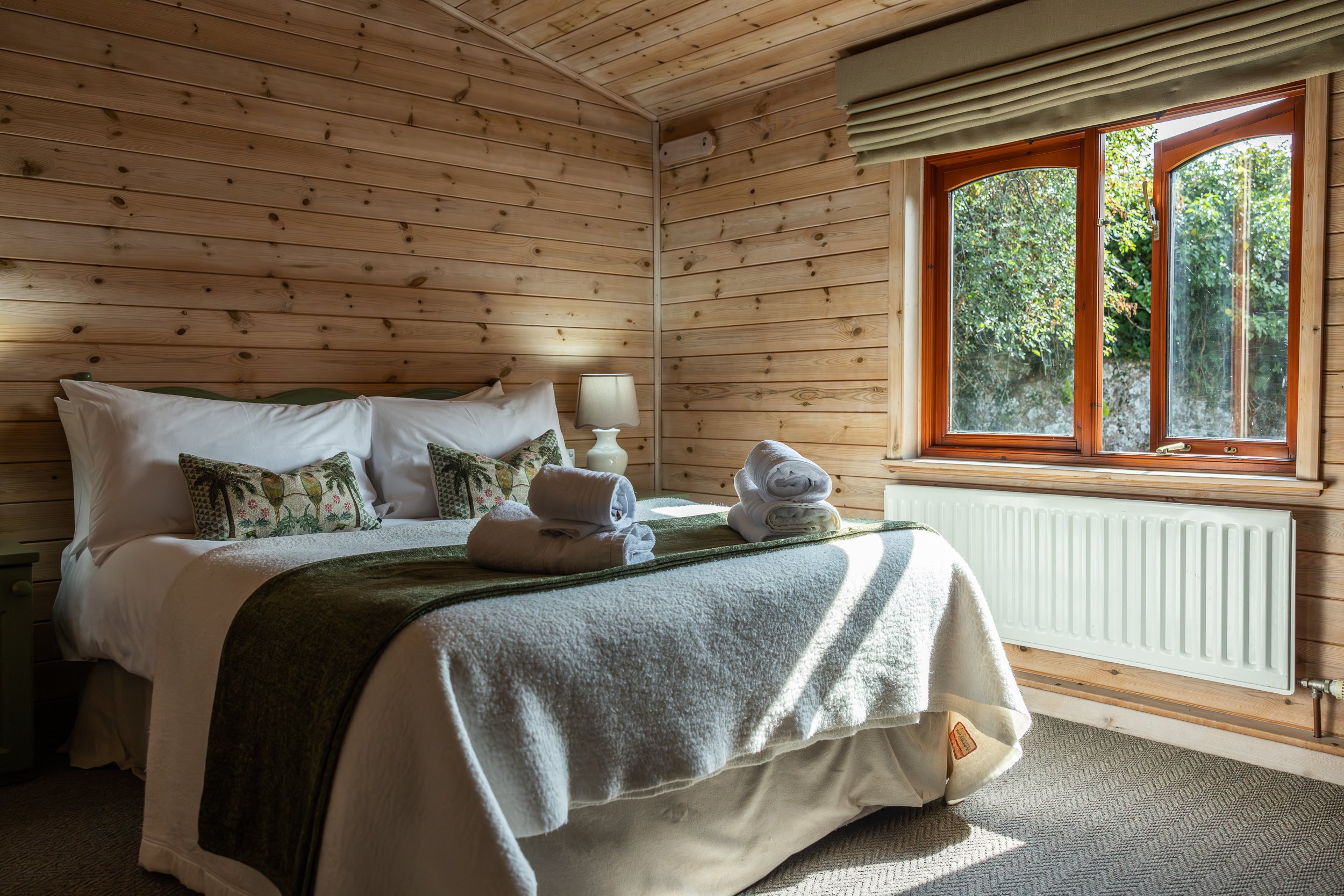 A cozy bedroom with wooden walls and a large window showing greenery outside. The bed has white sheets, decorative pillows with bird patterns, and rolled towels on top. A nightstand with a lamp is beside the bed.