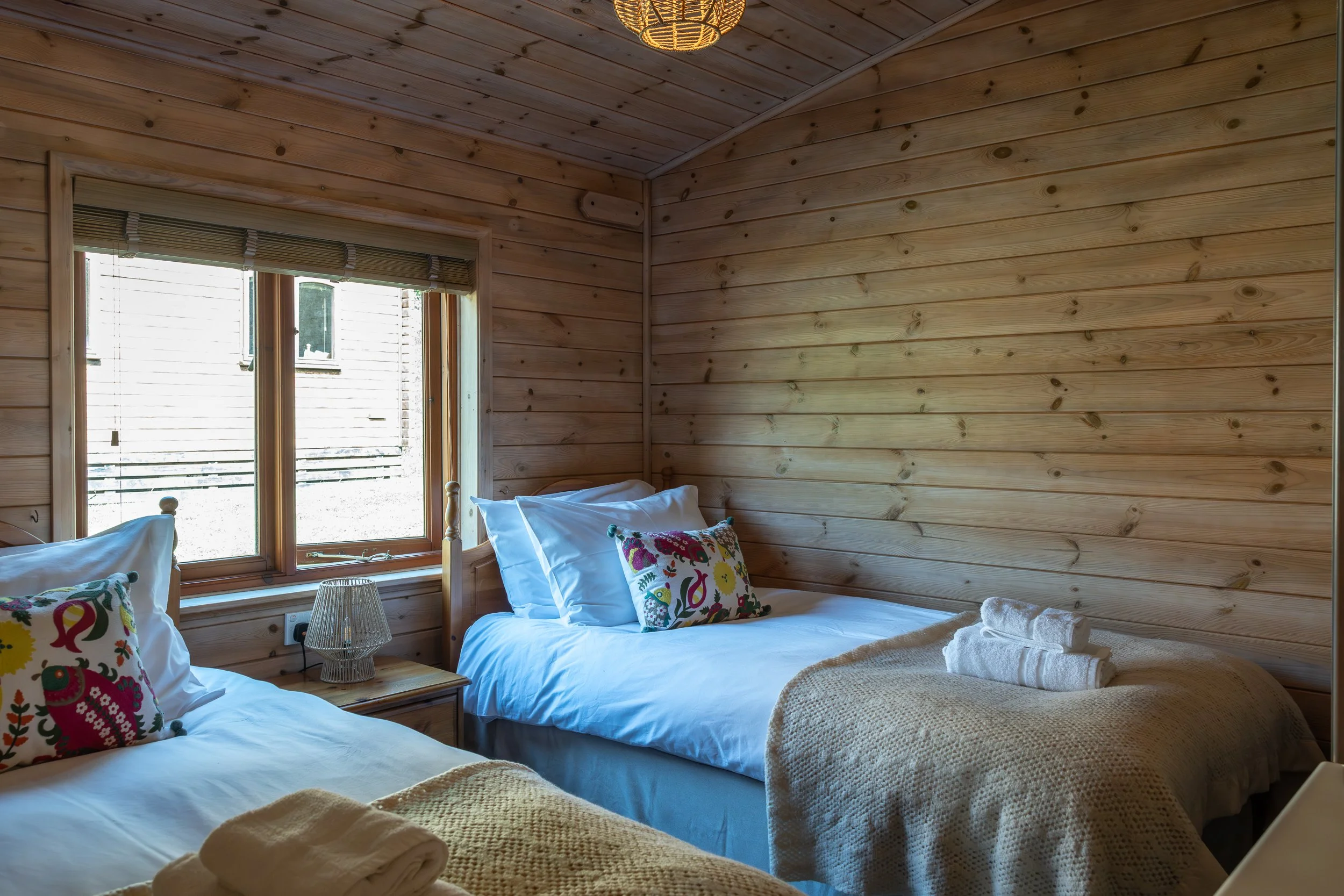 Two beds in a cozy wooden bedroom with pillows and rolled towels, next to a window with blinds, featuring a side table and lamps.