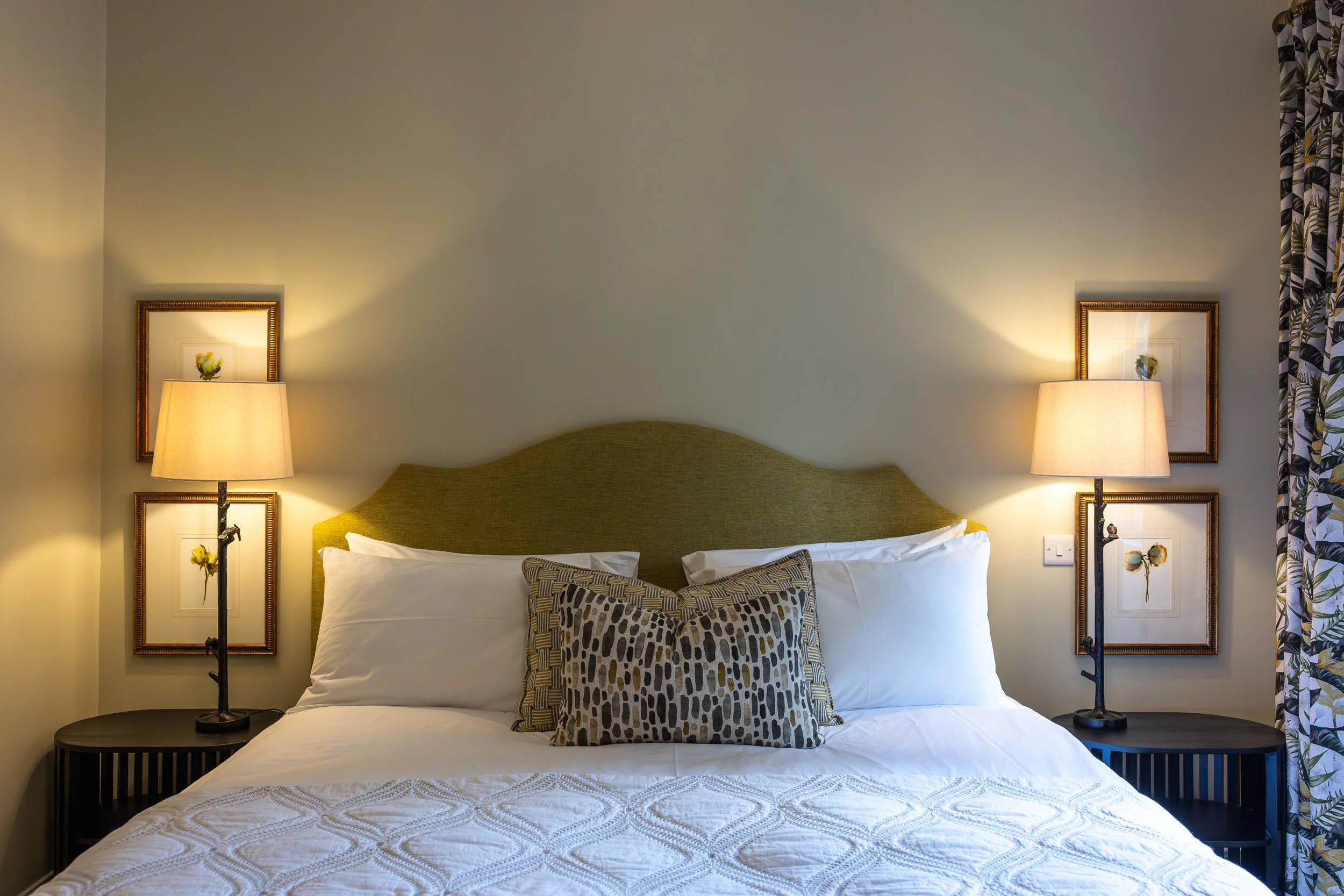 Twin beds with white bedding and patterned pillows, a green headboard, two nightstands with lamps, framed floral artwork on the wall, and patterned curtains.