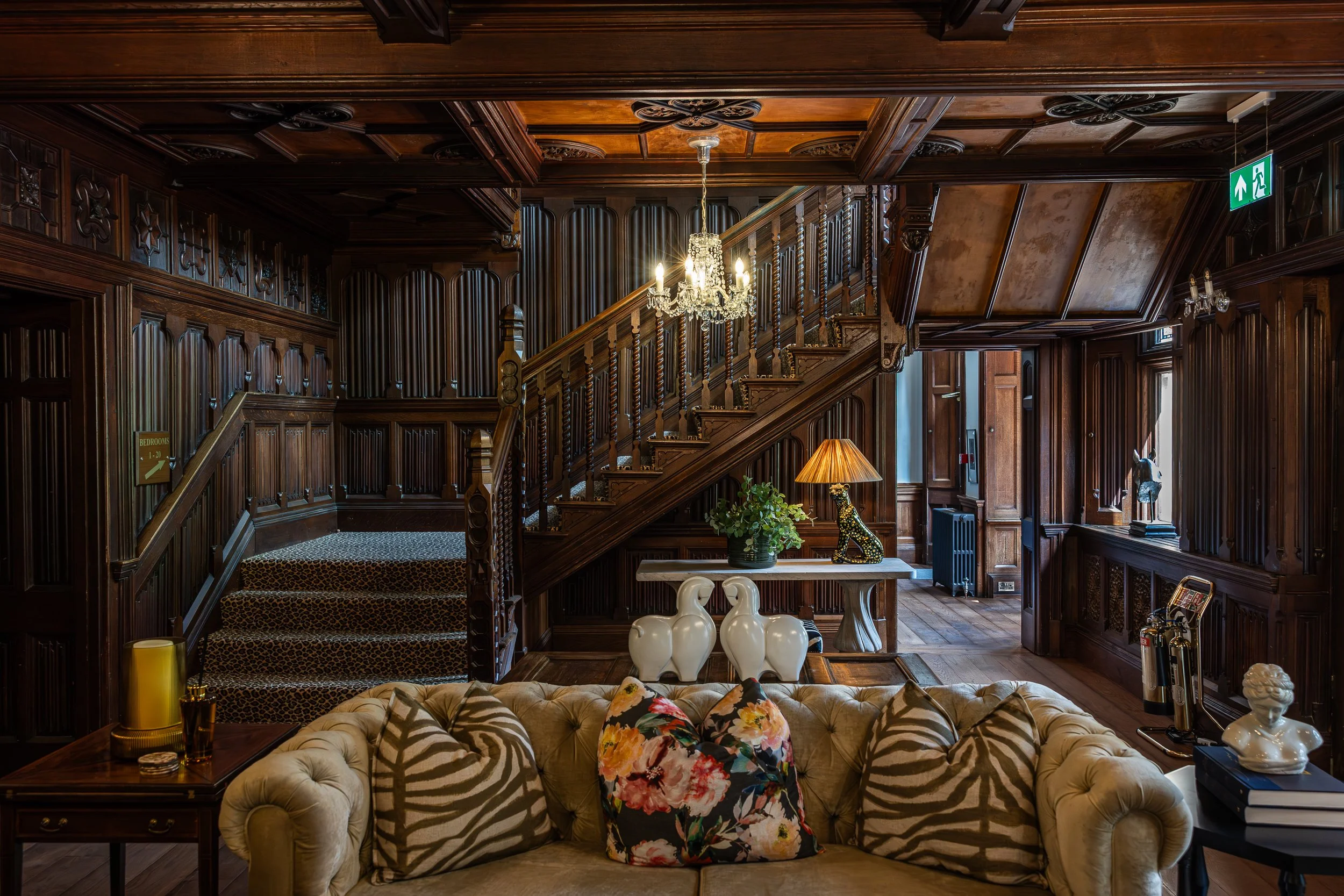 Elegant wooden interior with staircase, chandelier, decorative lamps, and upholstered furniture with floral and zebra print pillows.
