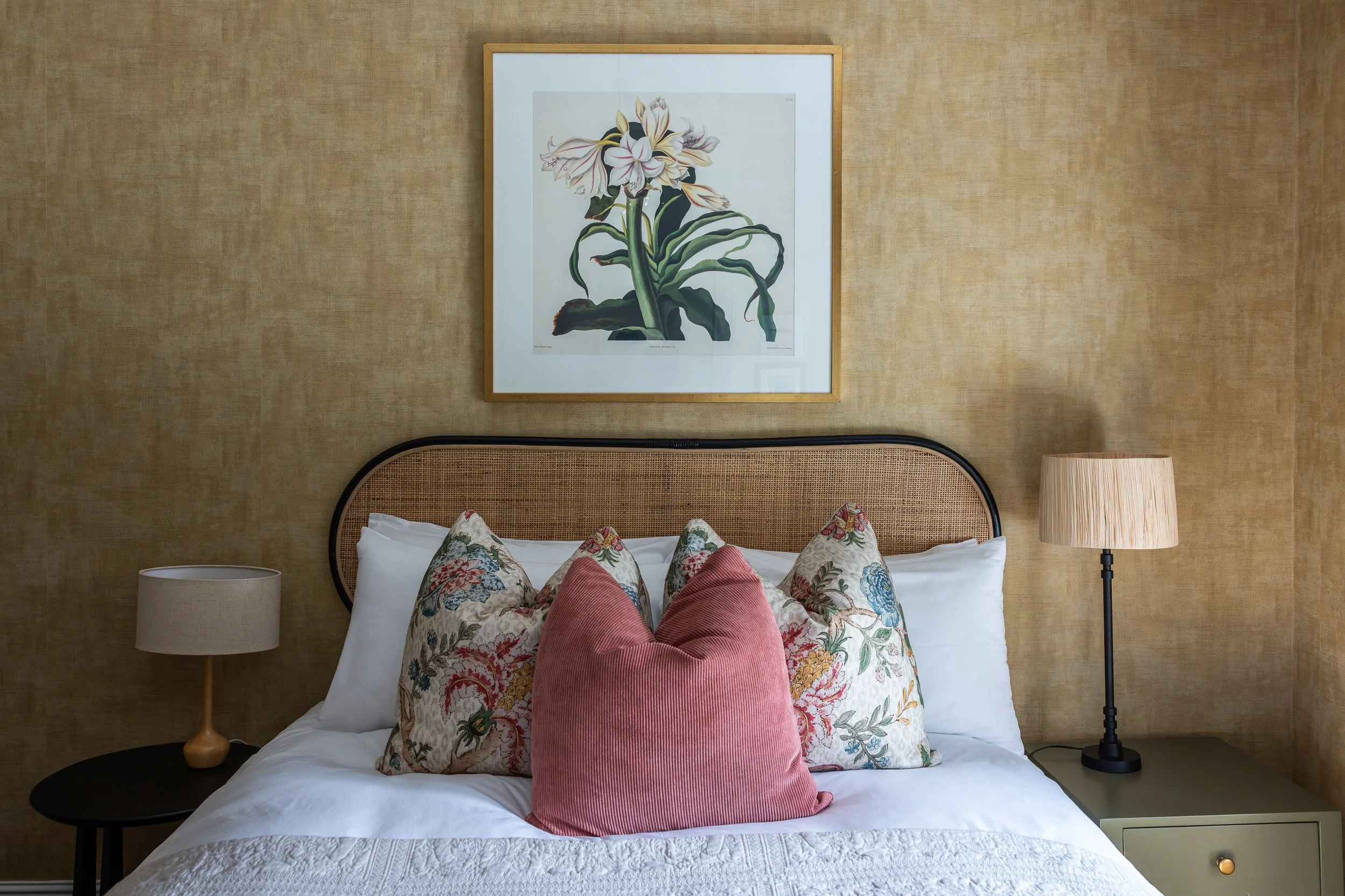 A bedroom with a bed featuring floral and velvety pink pillows, a framed floral artwork above the headboard, beige walls, and two bedside lamps, one beige and one black with beige shade.