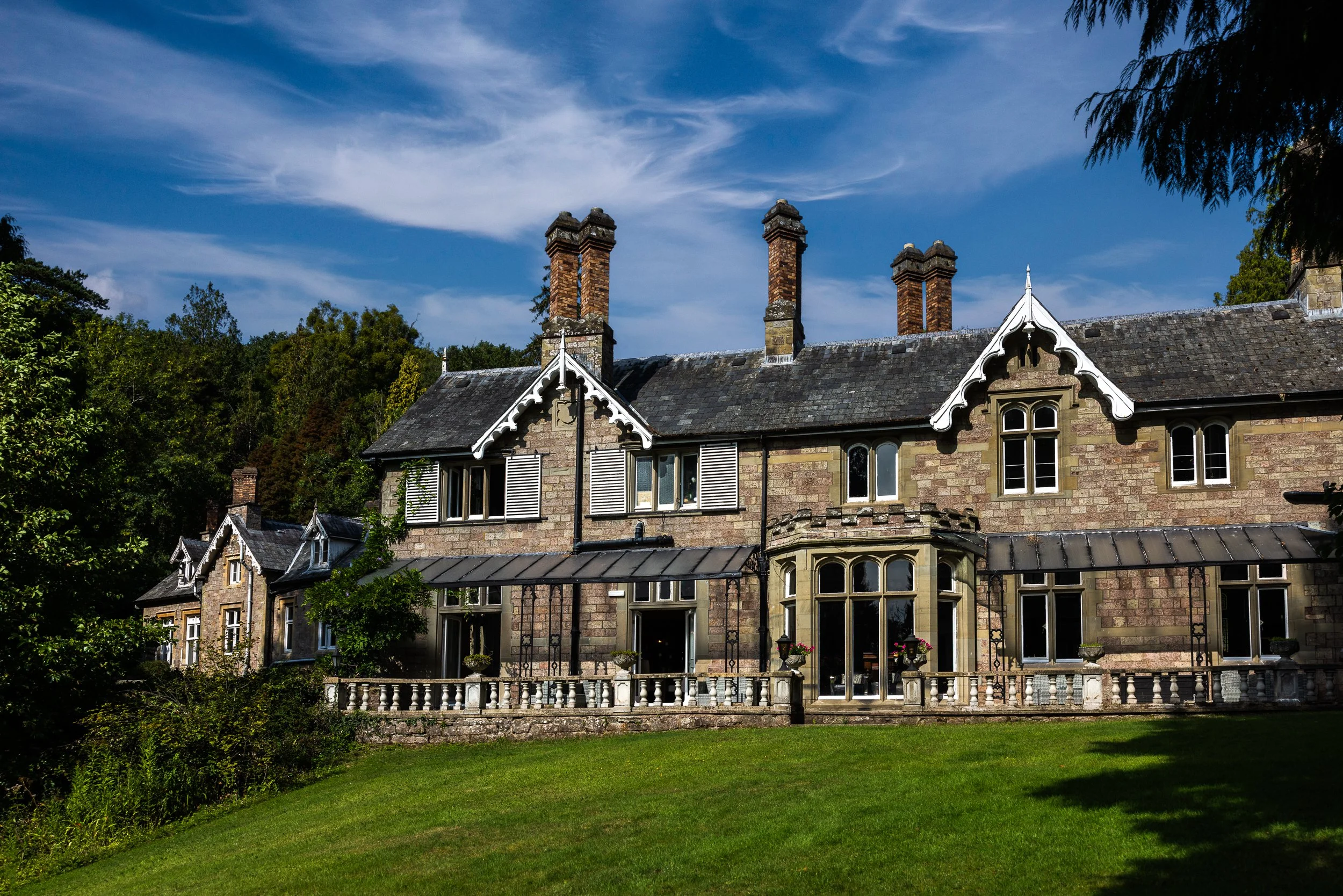 A large stone house with multiple chimneys and decorative white trim, surrounded by green trees and a well-kept lawn under a blue sky with wispy clouds.