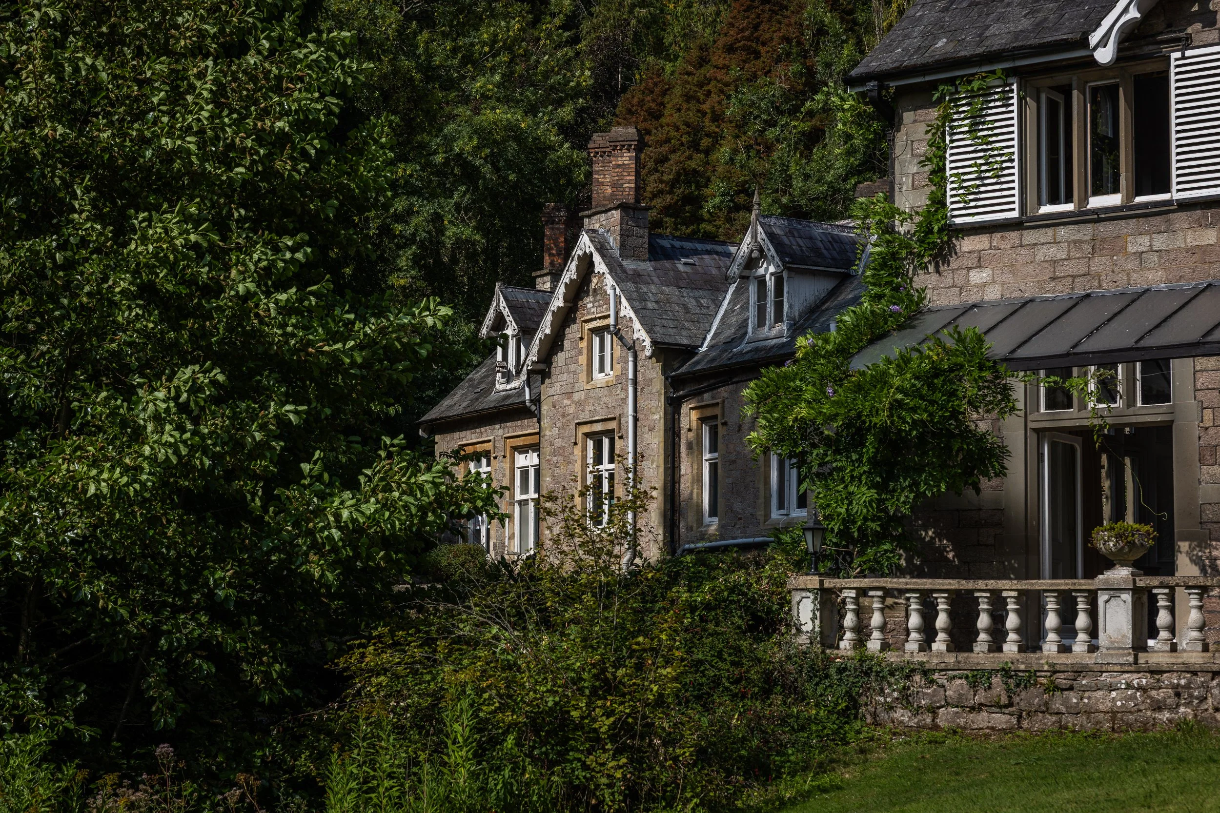 A stone house with white windows, surrounded by green trees and bushes.