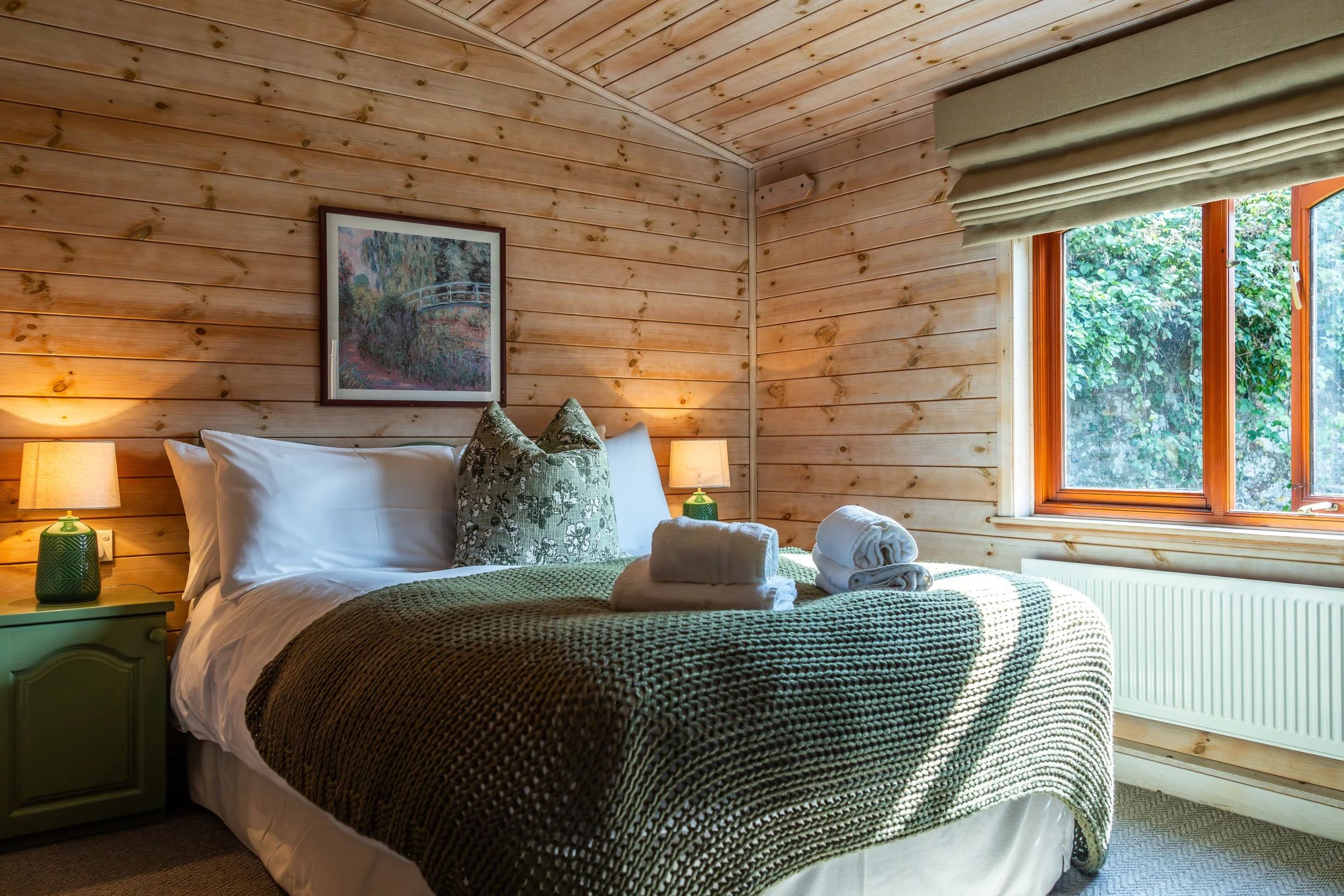 Cozy bedroom with wood-paneled walls, a bed with white pillows and a green knitted blanket, two rolled white towels on the bed, a green nightstand with a lamp, a window showing greenery outside, and a framed artwork on the wall.