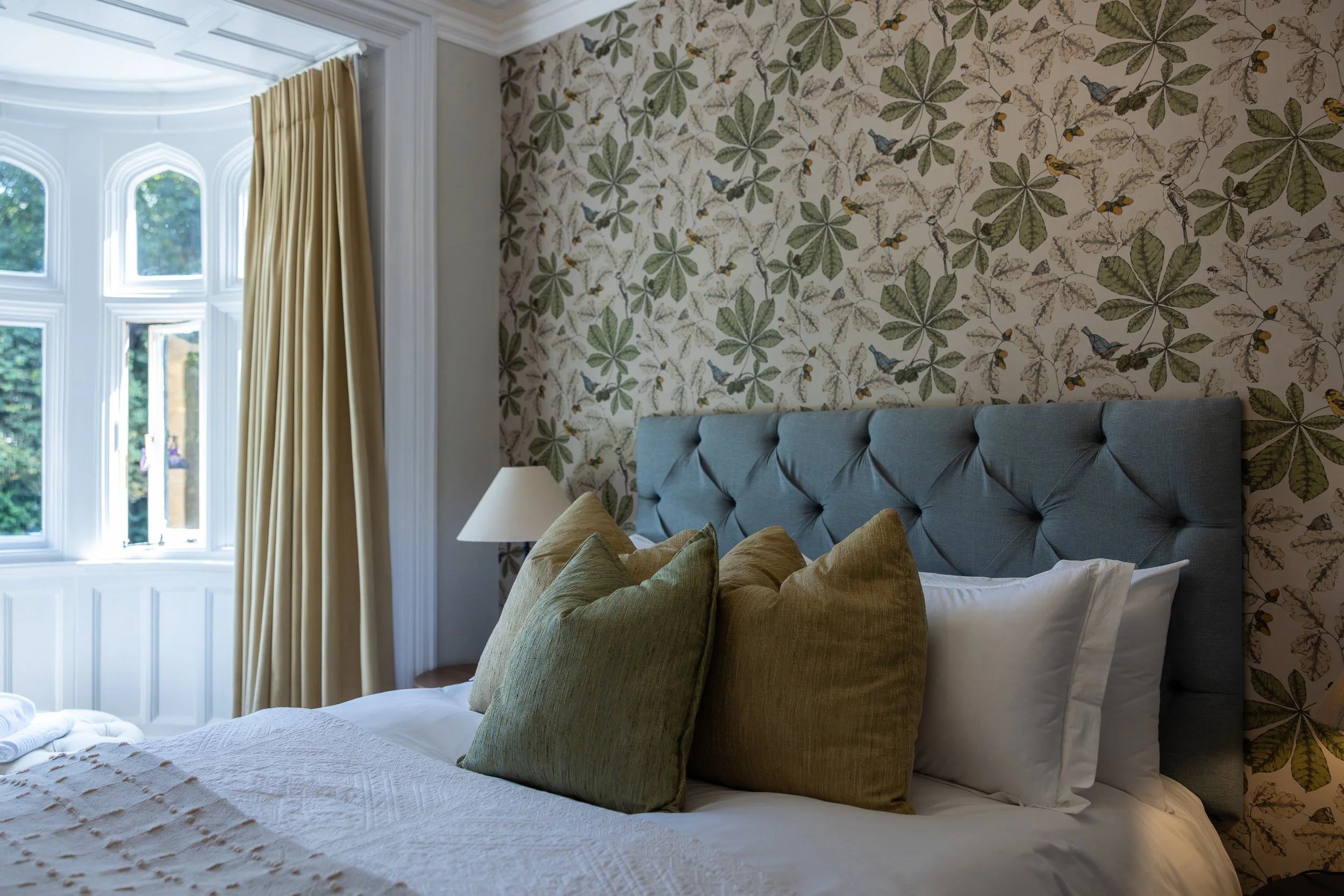 A bedroom with a large window with cream-colored curtains, a patterned wallpaper with leaves and birds, a gray upholstered headboard, white bedding, and multiple beige pillows, with a white lamp on a bedside table.