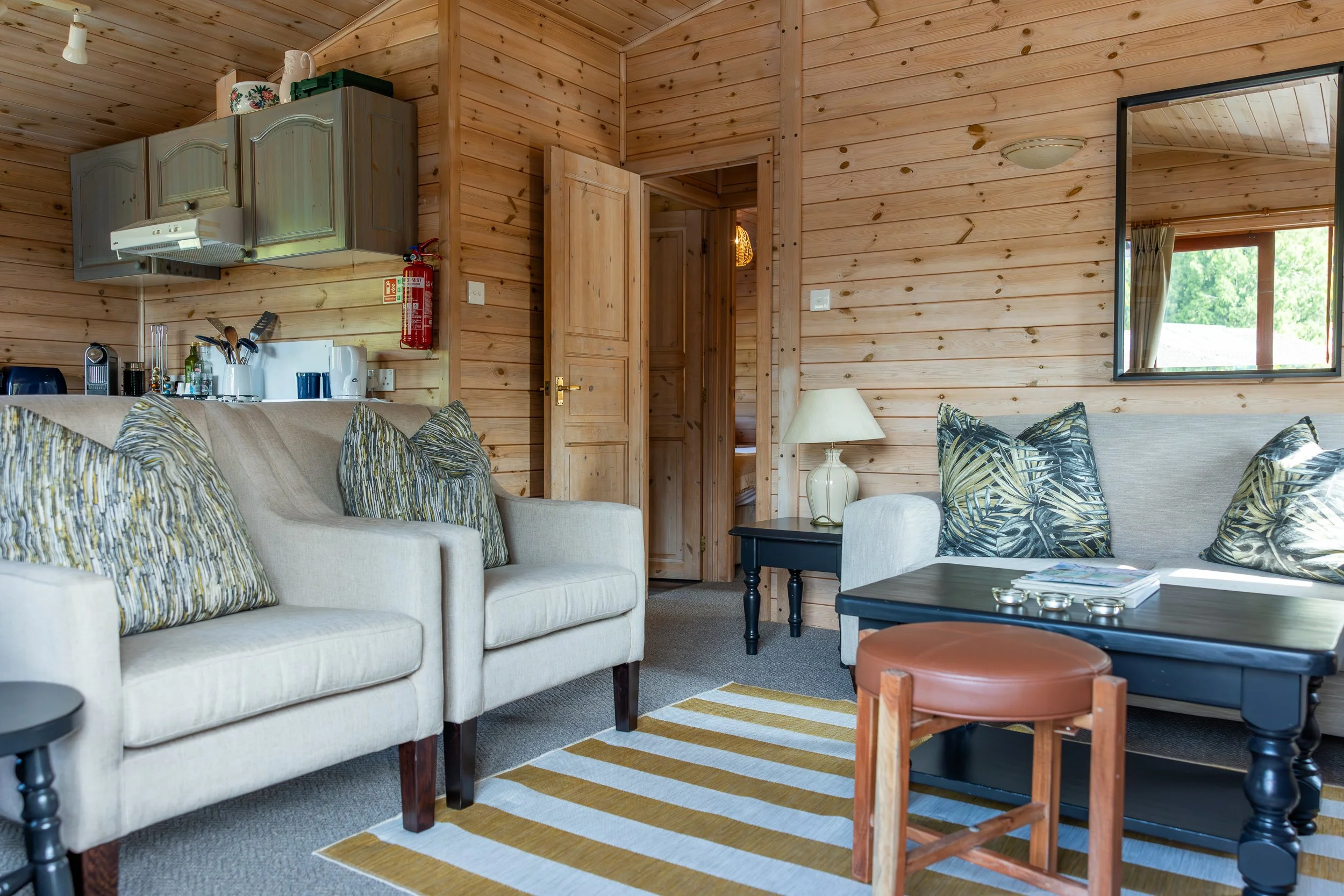 Living room with cream sofas, throw pillows, black coffee table, side table, lamp, striped yellow and white rug, wooden walls and ceiling, kitchen area with gray cabinets, kitchen countertop with appliances, and window showing greenery outside.