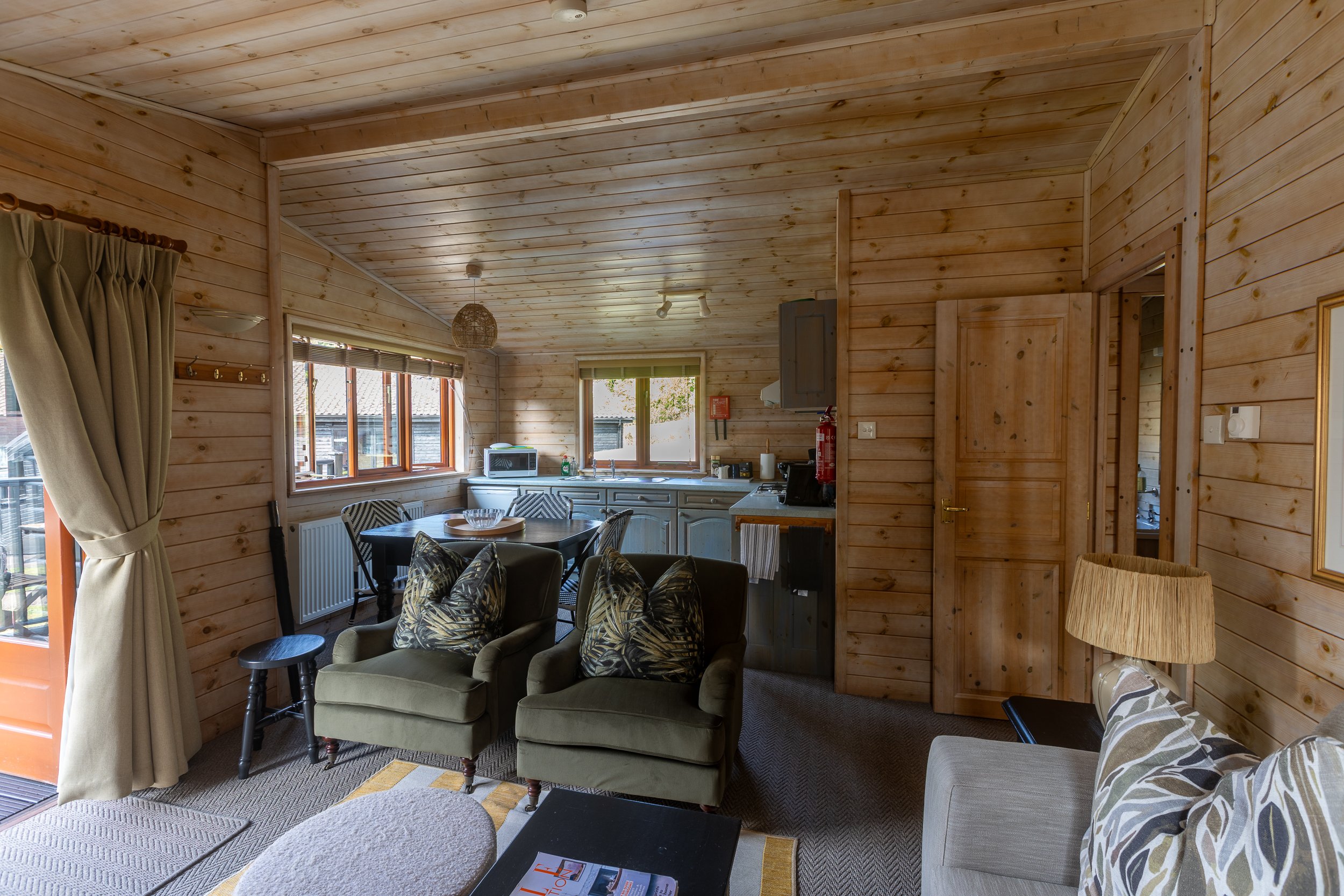 Interior of a cozy wooden cottage with living area, armchairs, sofa, dining table, kitchen area with cabinets, window, and door.