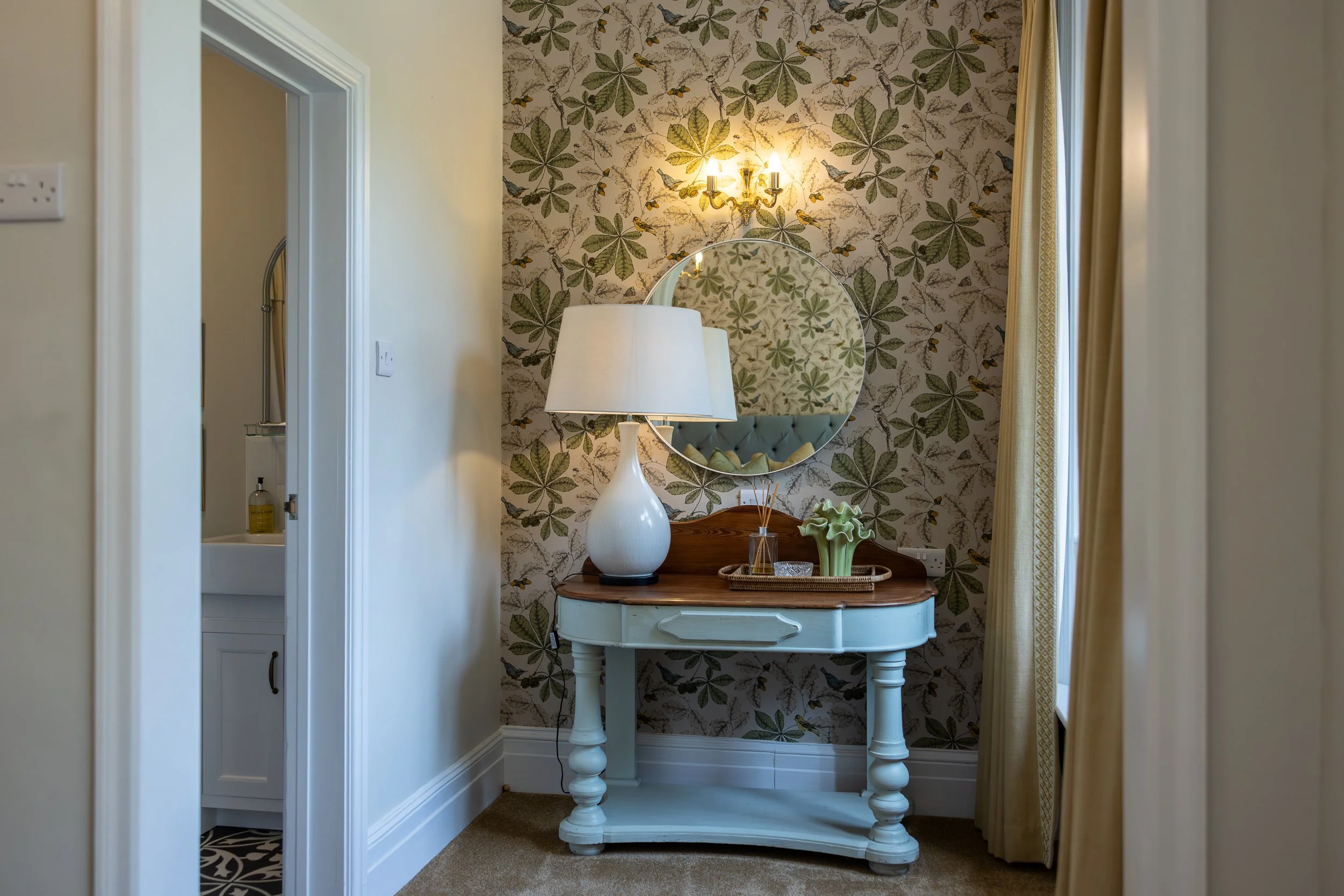 A vintage white and wood vanity table with a large white lamp, a small vase with reed diffuser, and a green ceramic flower sculpture, placed against a floral wallpapered wall with green leaves and yellow flowers, beneath a round mirror and a wall sco