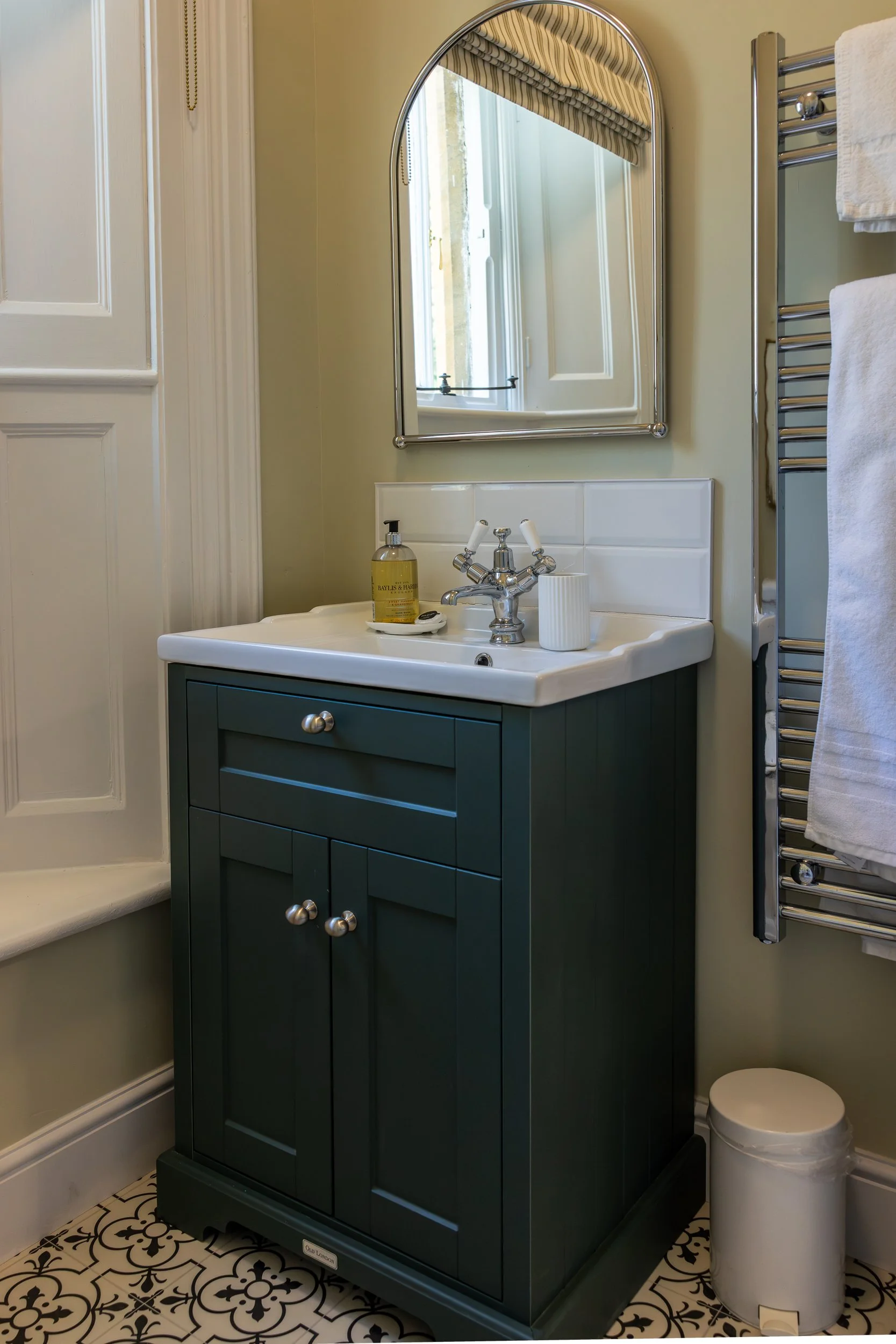 Small bathroom vanity with teal cabinet, white ceramic sink, silver hardware, mirror, soap dispenser, toothbrush holder, and towels, adjacent to window with partial view of interior and patterned floor tiles.
