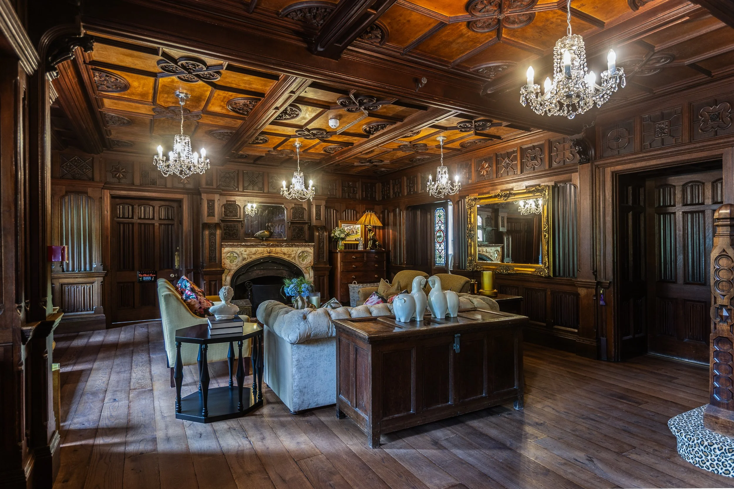 A luxurious living room with dark wood paneling, ornate ceiling, and multiple chandeliers. There are sofas, a fireplace, a large mirror, and decorative items.