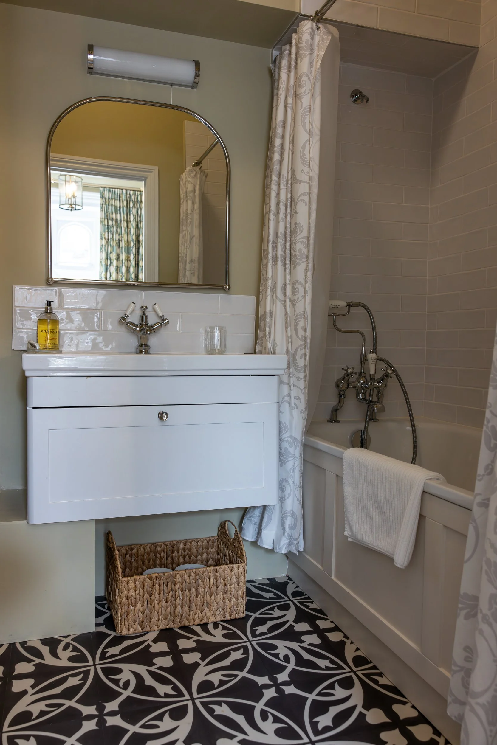 A bathroom with a white vanity, an oval mirror, a wall-mounted light fixture, a bathtub with a shower curtain, and patterned black and white floor tiles.