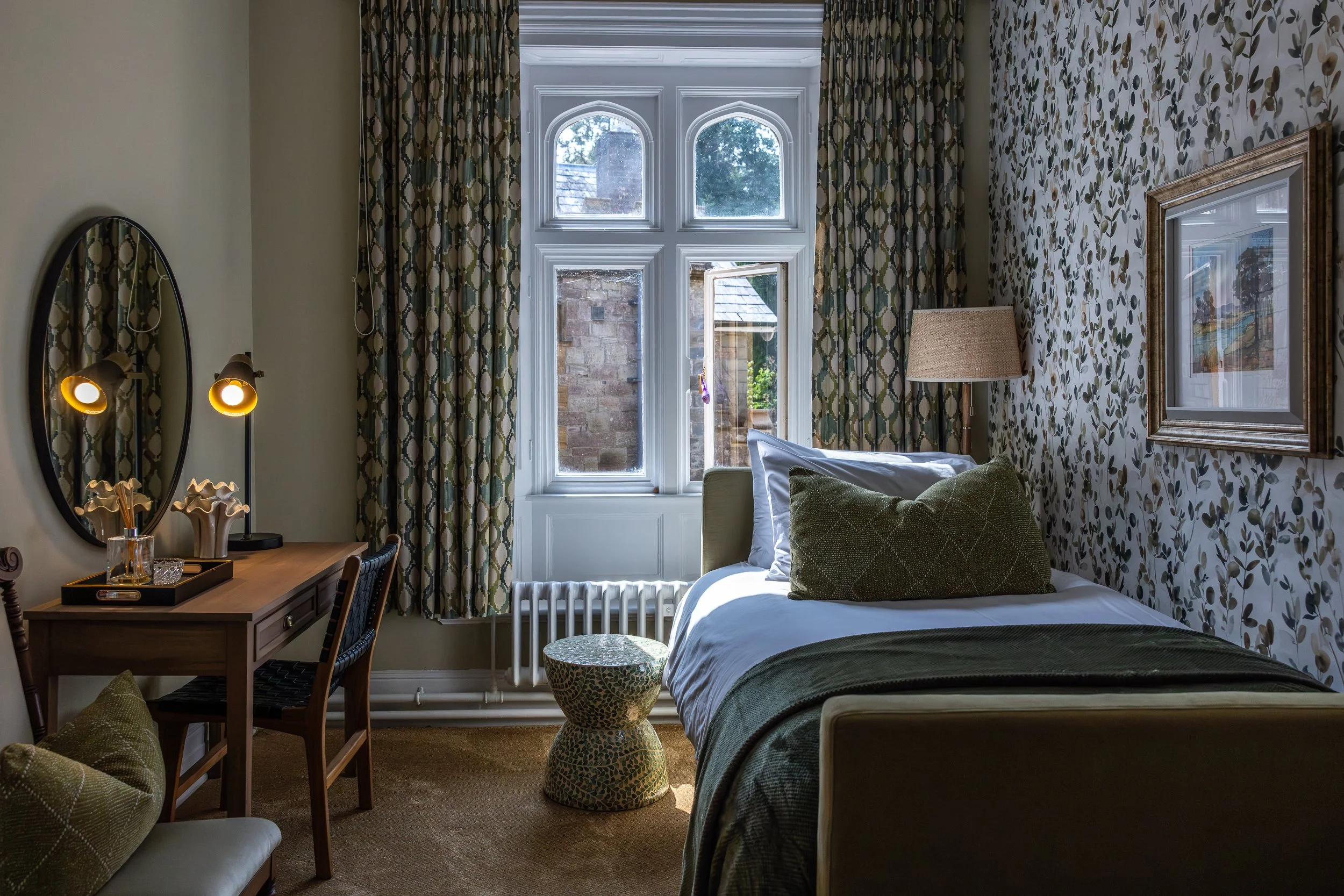 A cozy bedroom with a bed, green pillows, and a floral wallpaper. A window with patterned curtains lets in natural light. There is a wooden desk with a mirror, lamps, and decorative items, and an upholstered stool beside the bed.