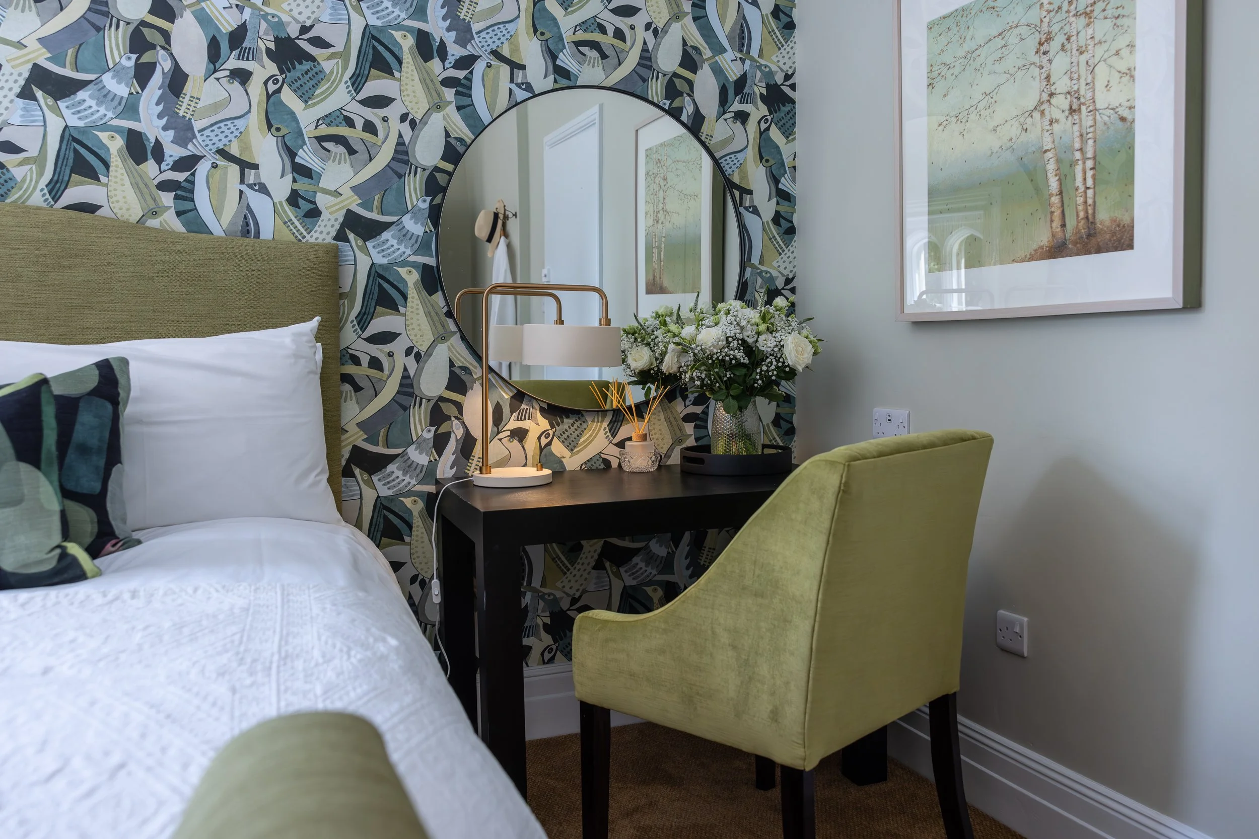 A bedroom corner with a bed, a black desk with a white lamp, a floral arrangement, a large round mirror, and an armchair. The wall behind the mirror features bird-patterned wallpaper, and there is a framed painting of a landscape on the adjacent wall