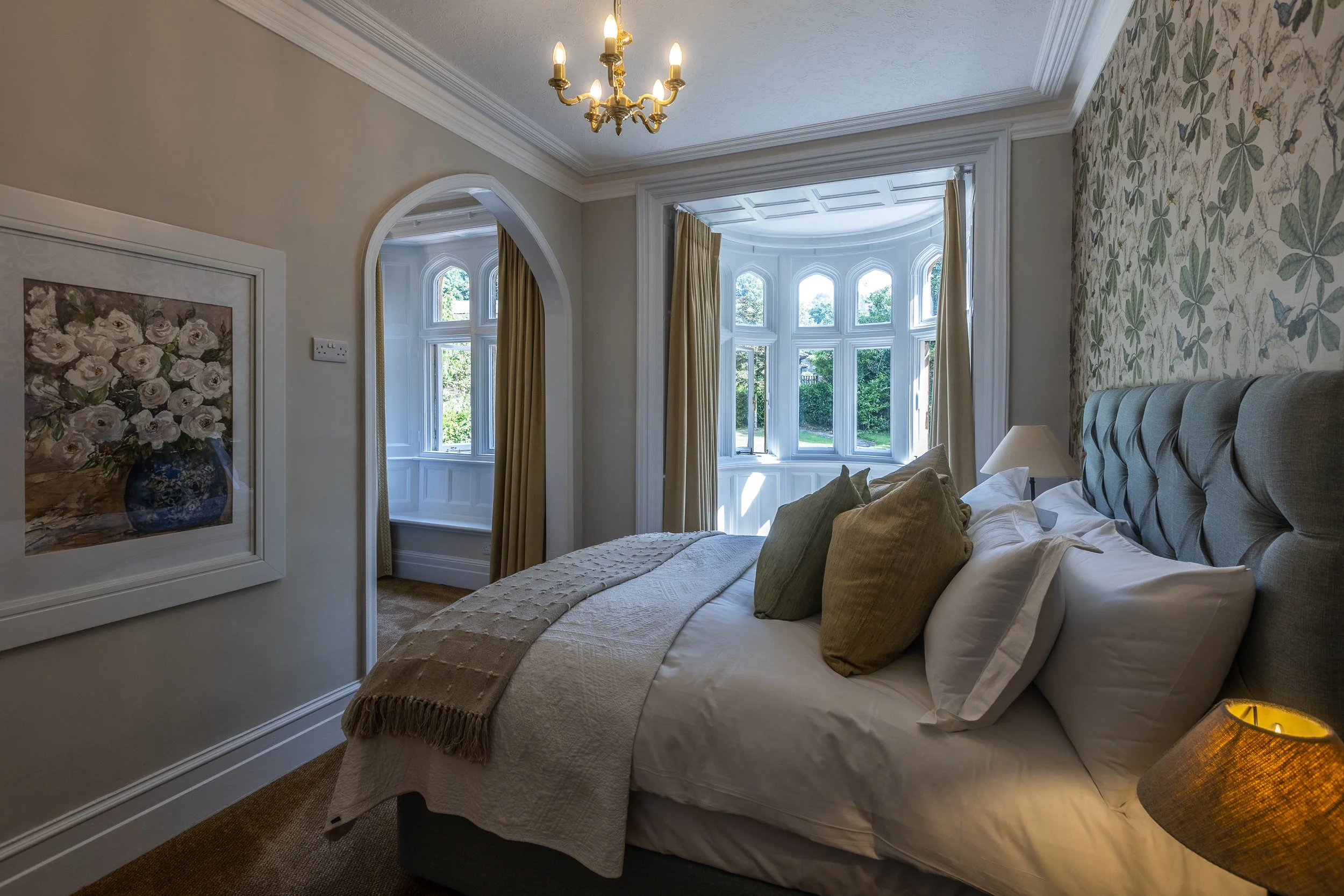 A cozy bedroom with a bed featuring green and white pillows, a beige throw blanket, a white quilt, and a fabric lampshade on the bedside table. The room has large bay windows with white trim, gold curtains, a floral wallpaper accent wall, a chandelie