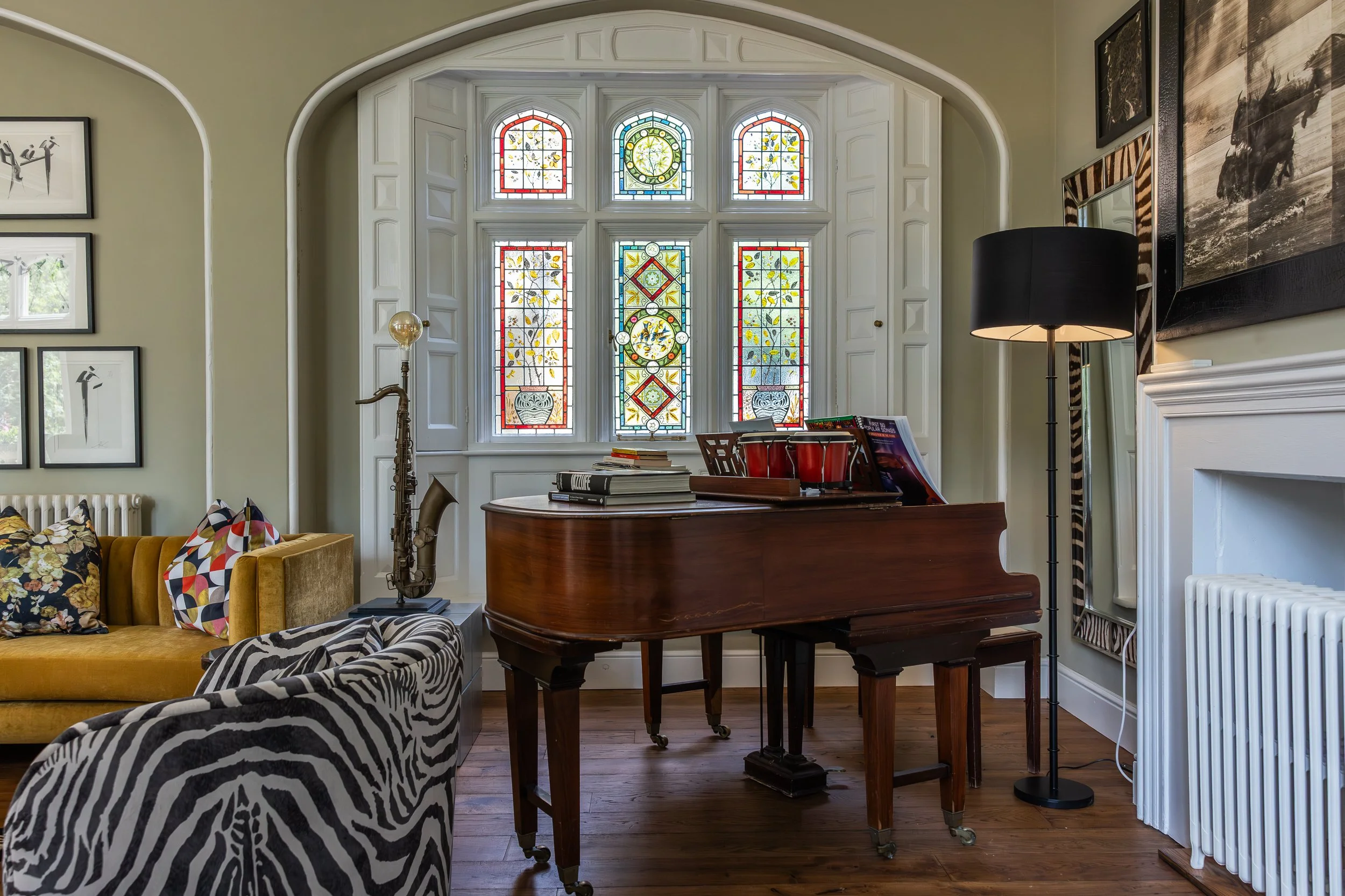 Living room with yellow sofa and zebra-patterned armchair, stained glass window, grand piano with books, saxophone, and lamp.