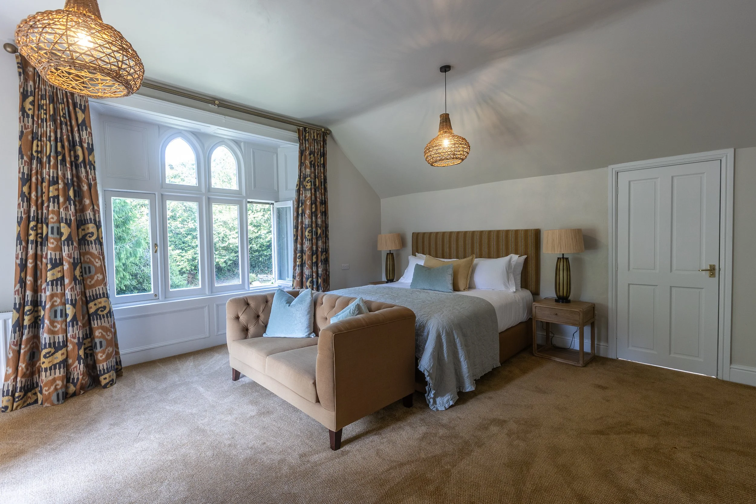A cozy bedroom with a large bed, a beige tufted sofa at the foot of the bed, and a window with patterned curtains
