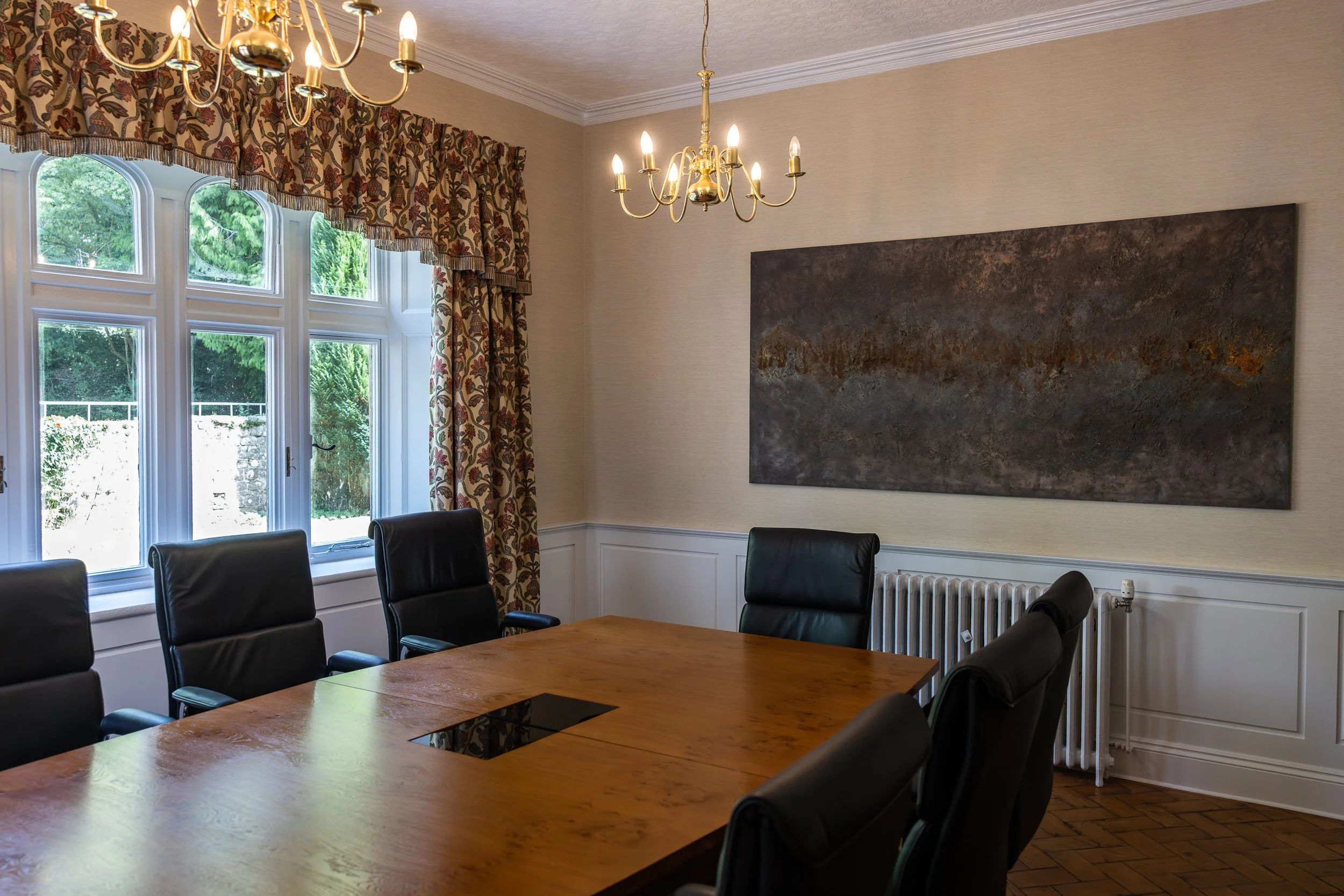 A formal conference room with a wooden table surrounded by black leather chairs, large window with floral curtains, two chandeliers, and an abstract painting on the wall.