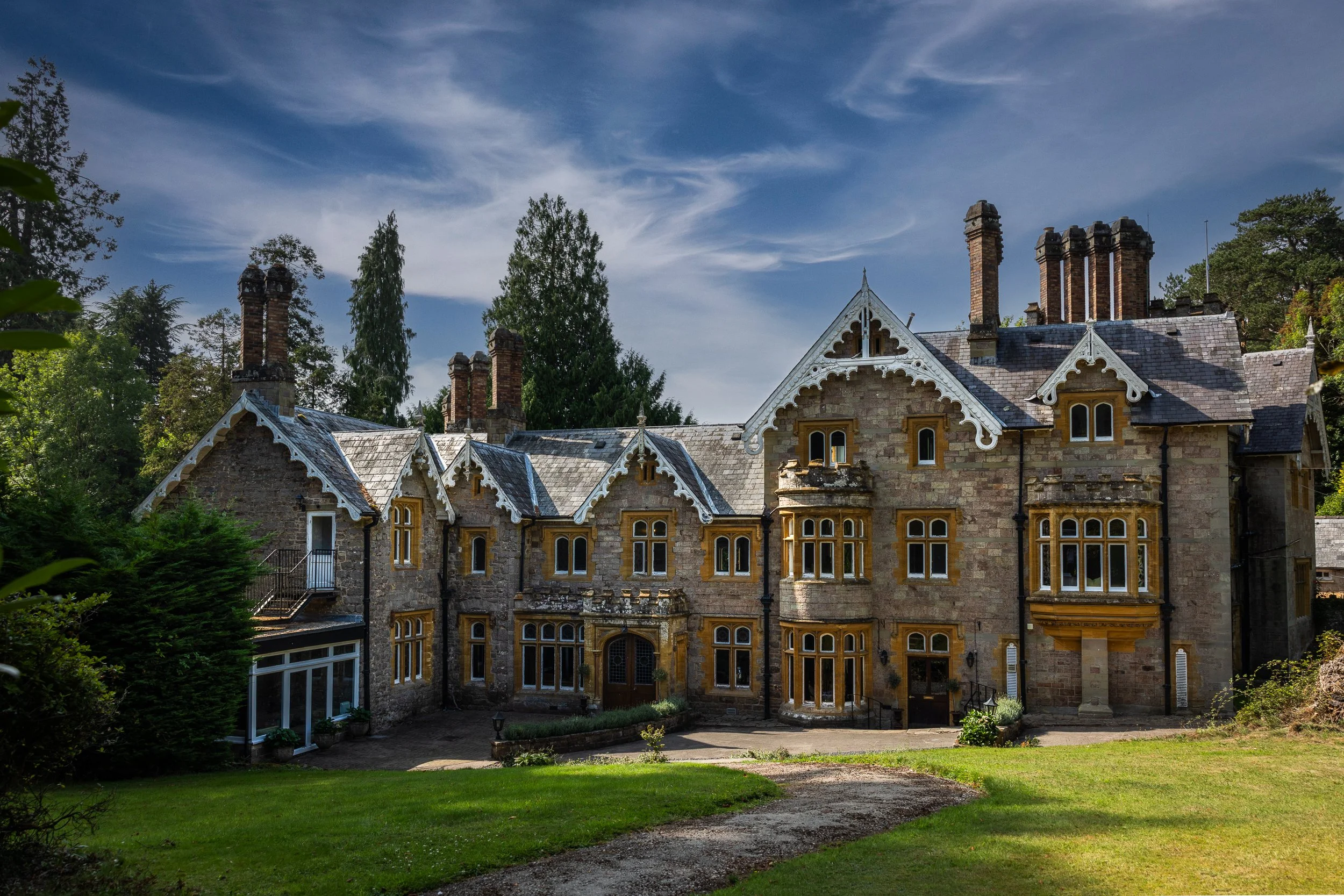 A large historic stone mansion with multiple chimneys, surrounded by green trees and a well-maintained lawn, under a blue sky with wispy clouds.