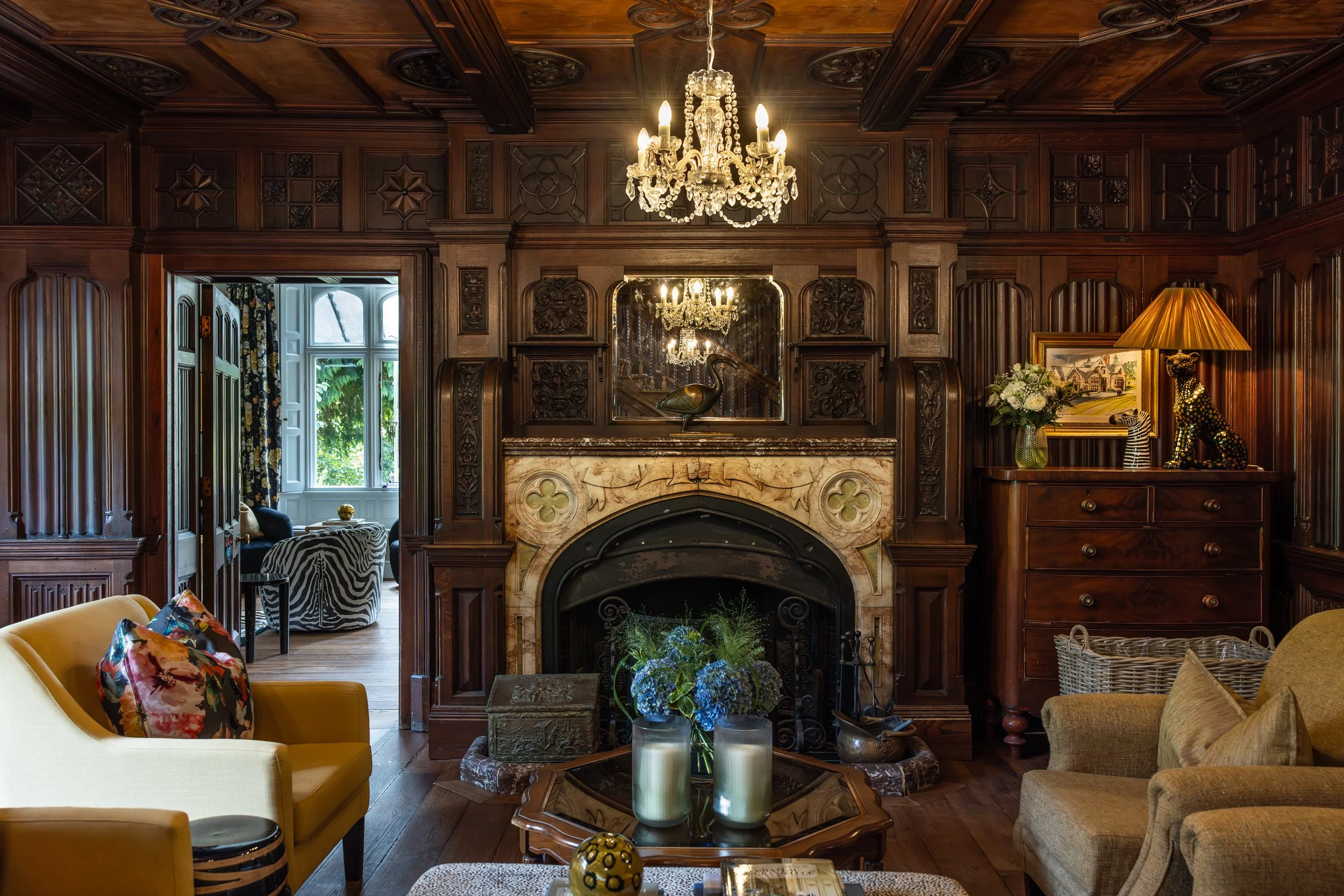 A vintage living room with a dark wood paneled wall, a marble fireplace, ceiling chandeliers, a mirror above the fireplace, a yellow sofa with a floral pillow, a beige armchair, a wooden coffee table with candles and flowers, a sideboard with a lamp,