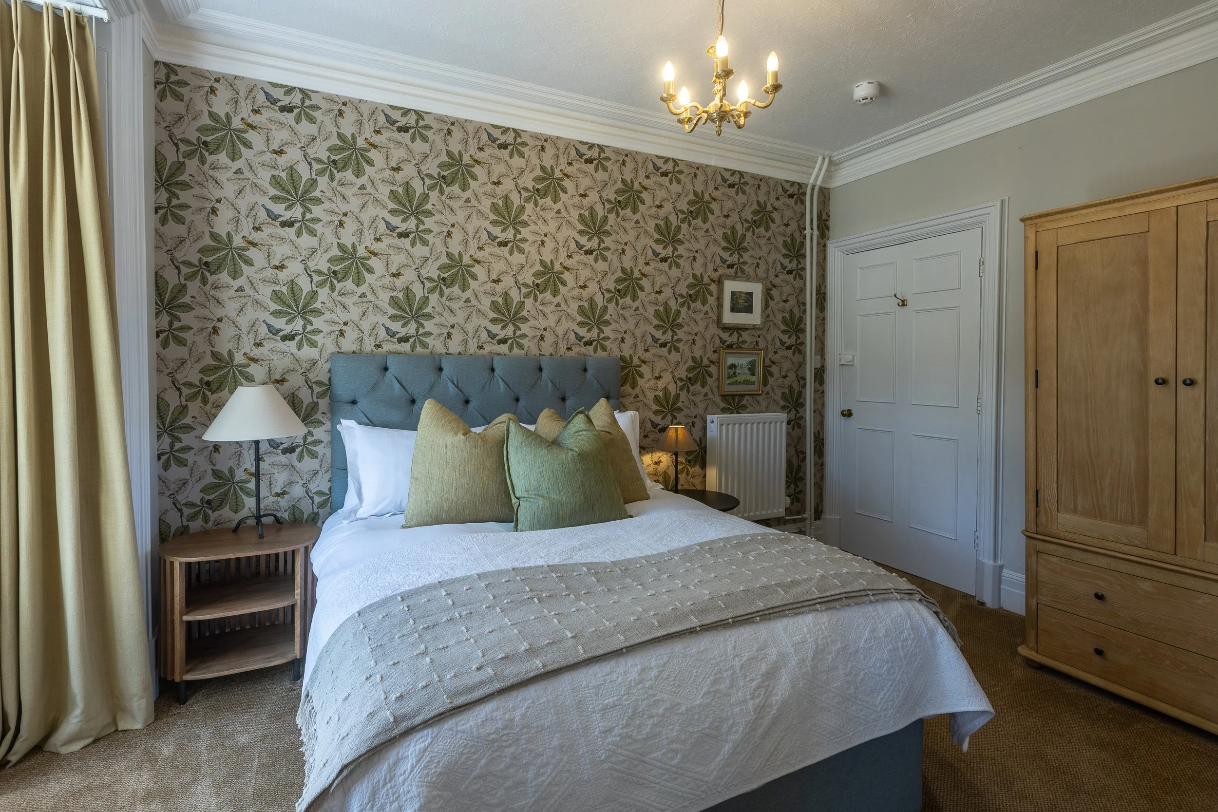 A neatly made bed with green pillows and a tufted blue headboard, two bedside tables with lamps, patterned wallpaper with leaves behind the bed, and a wooden wardrobe in a cozy bedroom with cream-colored curtains and decorative framed pictures on the
