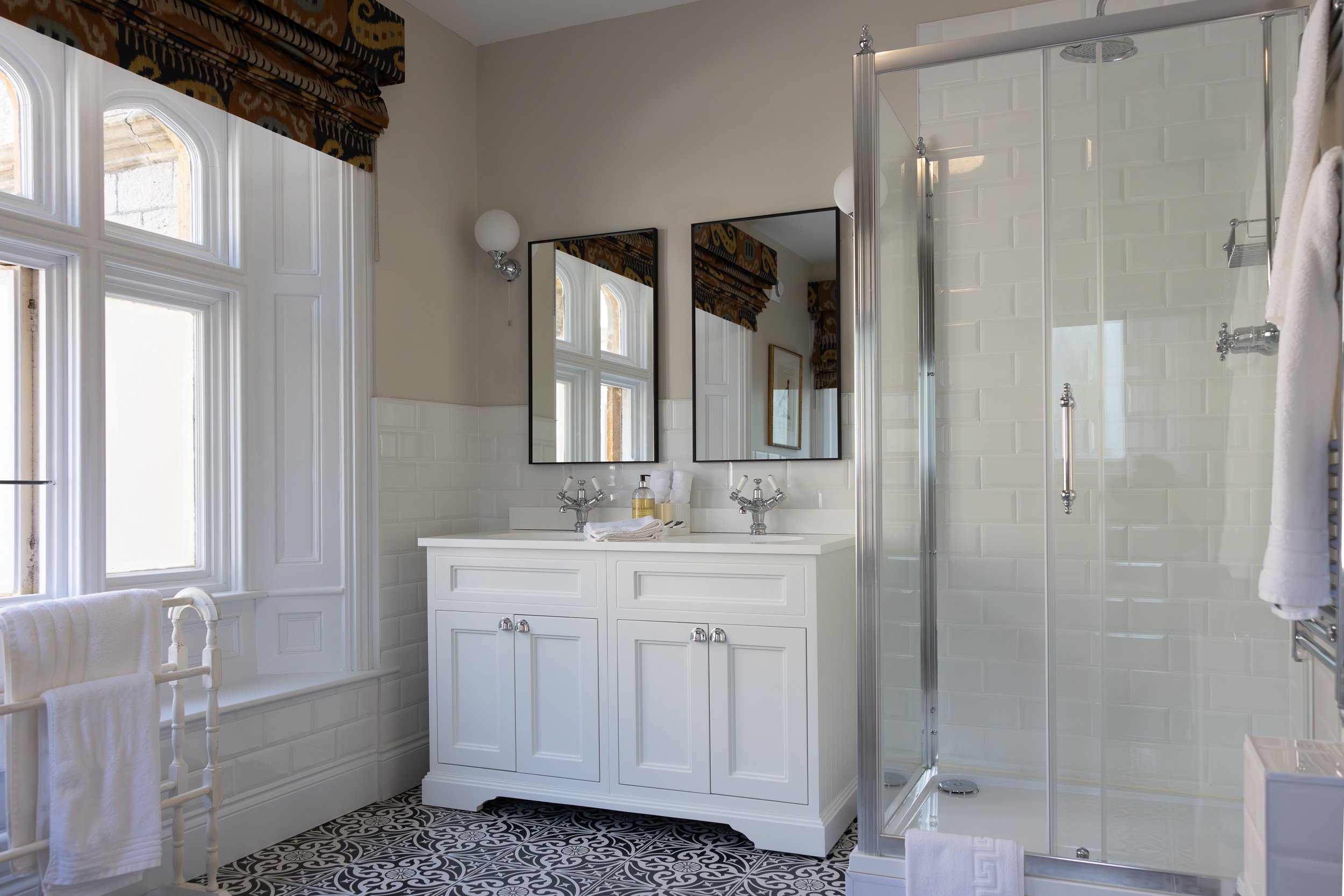 A bright bathroom featuring a white vanity with two sinks, two mirrors, a glass shower enclosure, patterned black-and-white floor tiles, white subway tile walls, a window with decorative curtains, and a towel rack with towels.