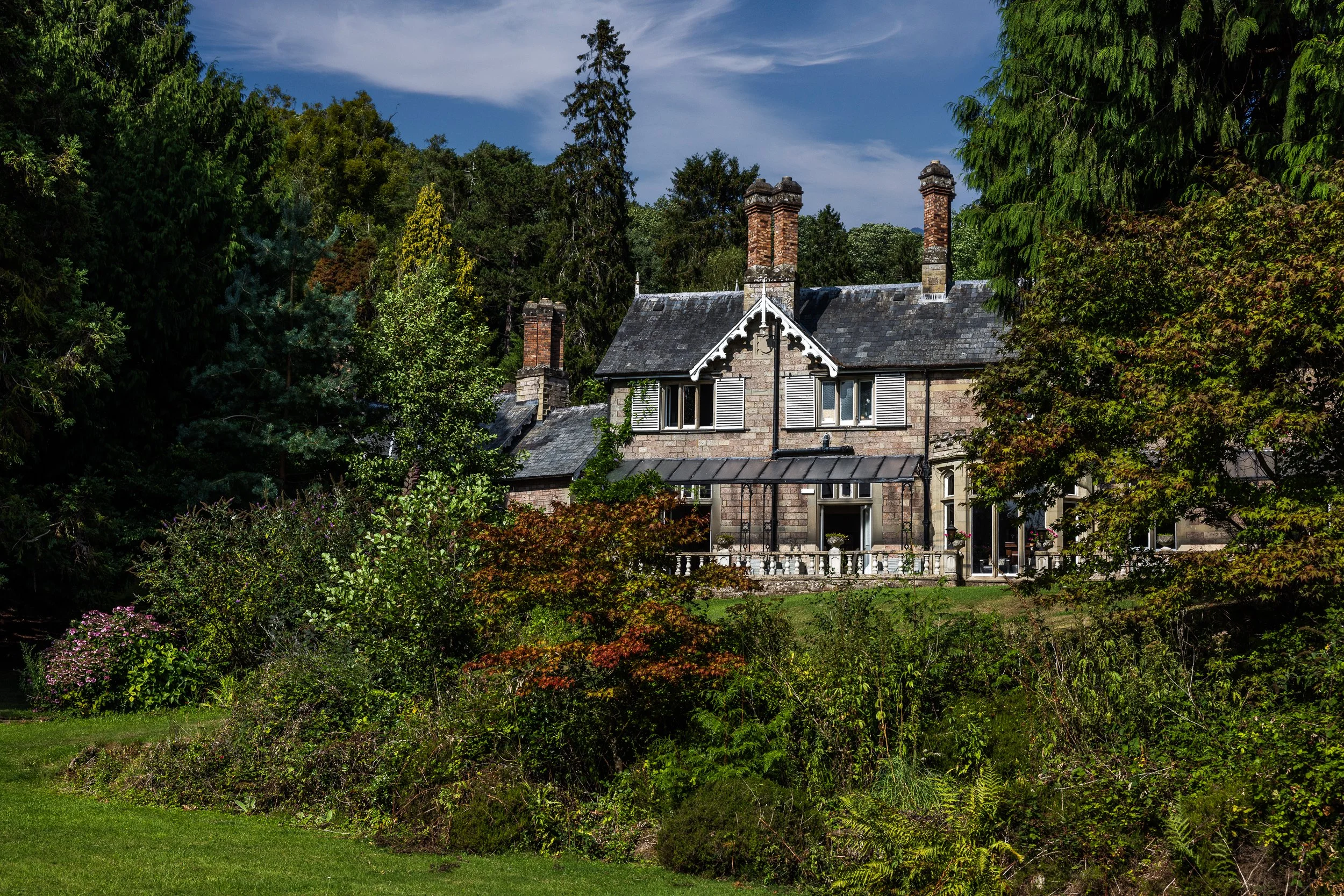 A large, historic house made of stone with multiple chimneys, surrounded by greenery and trees, under a partly cloudy sky.