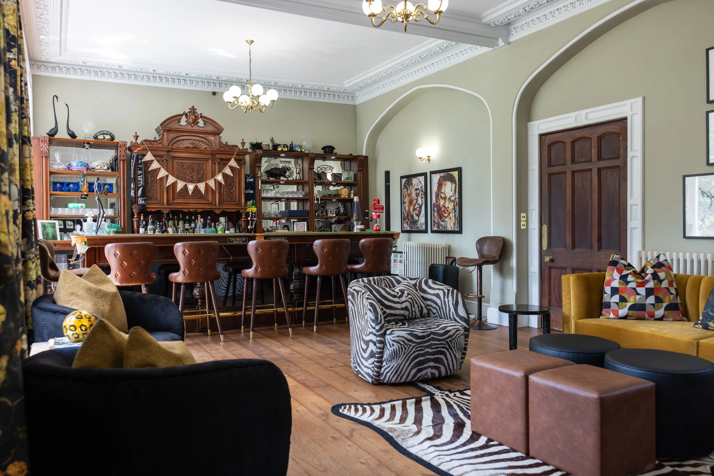 A cozy living room with a vintage feel, featuring a bar area with five brown leather bar stools, antique wooden shelving, artworks, and a mix of modern and retro furniture including a zebra print armchair, a yellow velvet sofa with patterned pillows,
