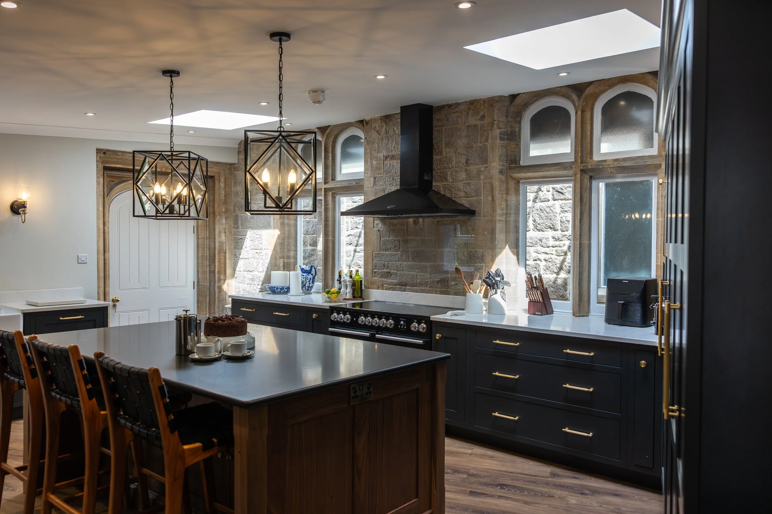 Modern kitchen with black cabinets, a large kitchen island, decorative pendant lights, stone walls, and multiple windows allowing natural light.