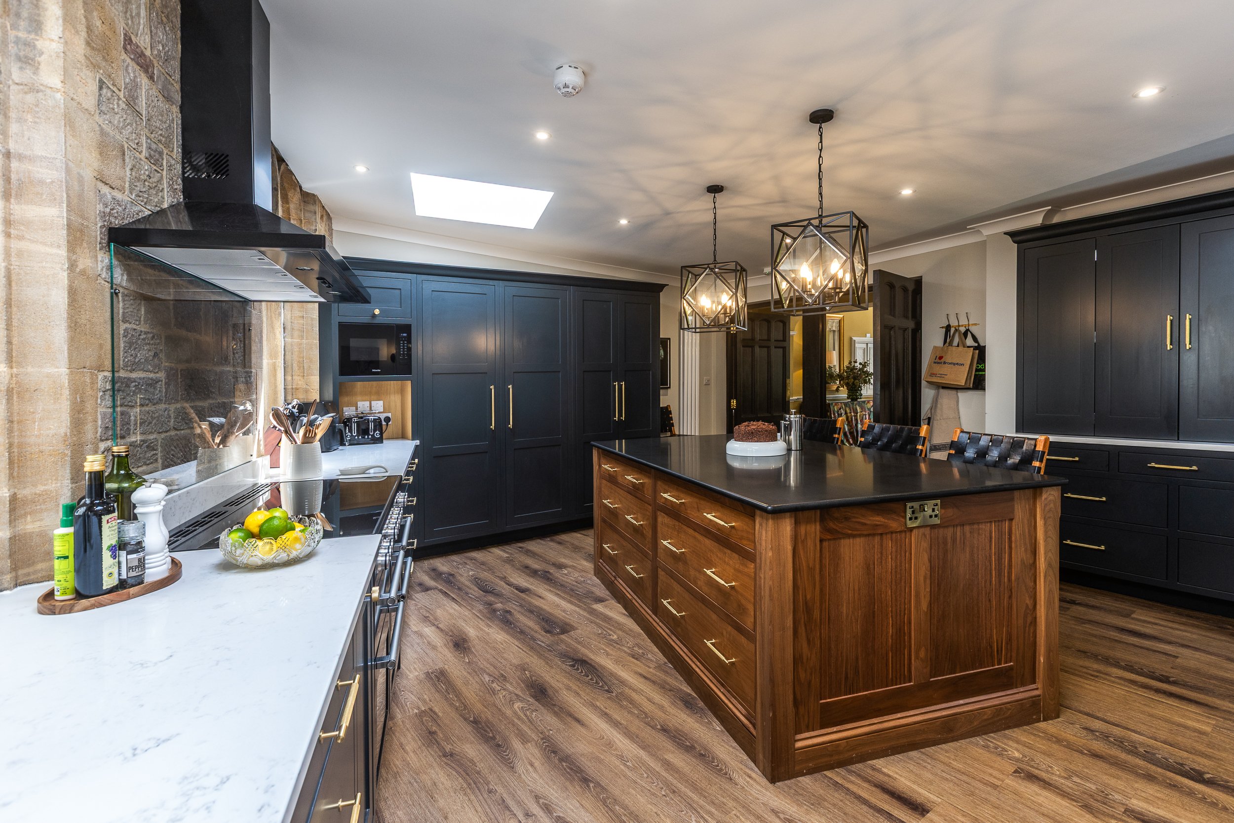 Modern kitchen with dark cabinets, a wooden island with a black countertop, and pendant lighting. The space features wood flooring, a white marble counter, and a brick wall near the stove.