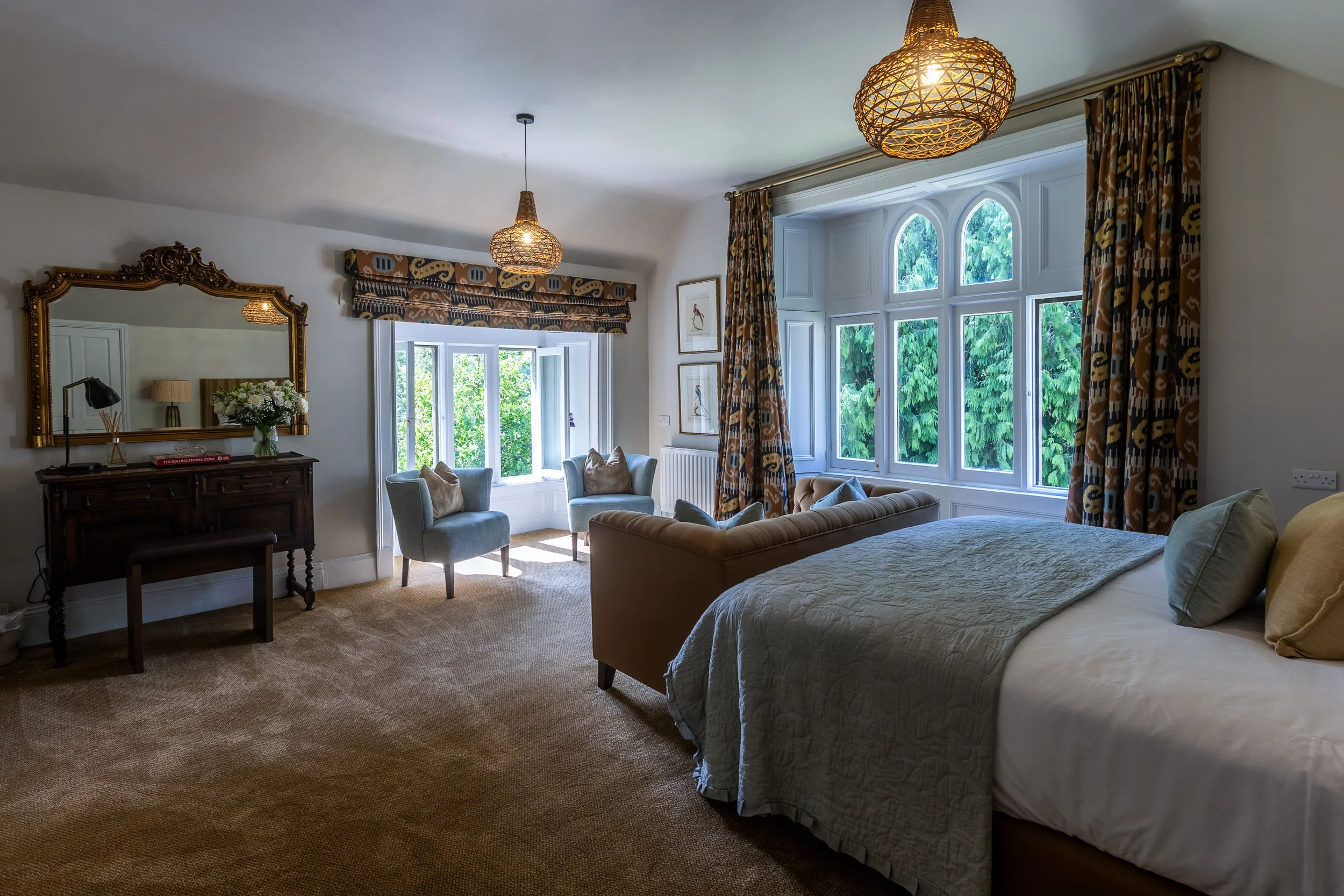 A bright bedroom with large bay windows, a bed with beige bedding, a curved beige sofa, two blue armchairs, a wooden dresser with a mirror, and patterned curtains.
