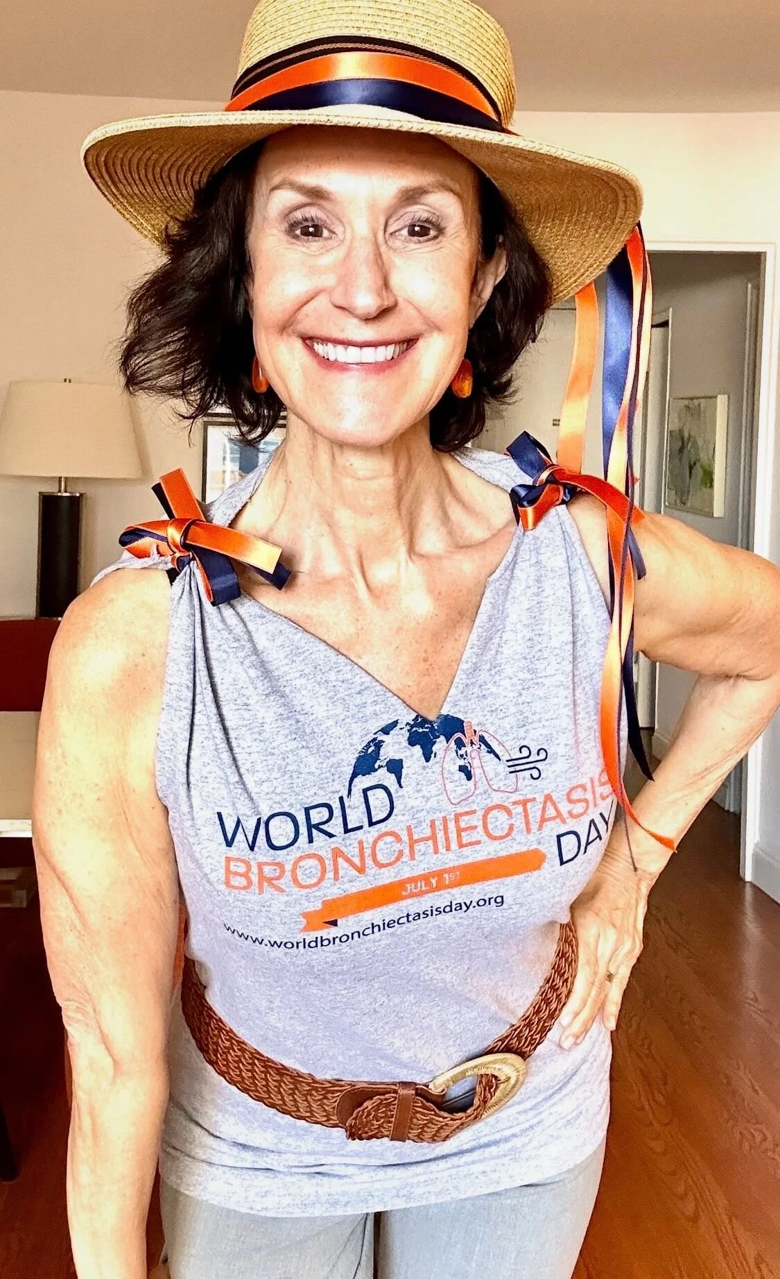 Linda Cooper Esposito, MPH wearing a wide-brimmed straw hat with colorful ribbons, a gray T-shirt with orange and blue text and graphics supporting World Bronchiectasis Day, a woven belt, and gray pants, standing